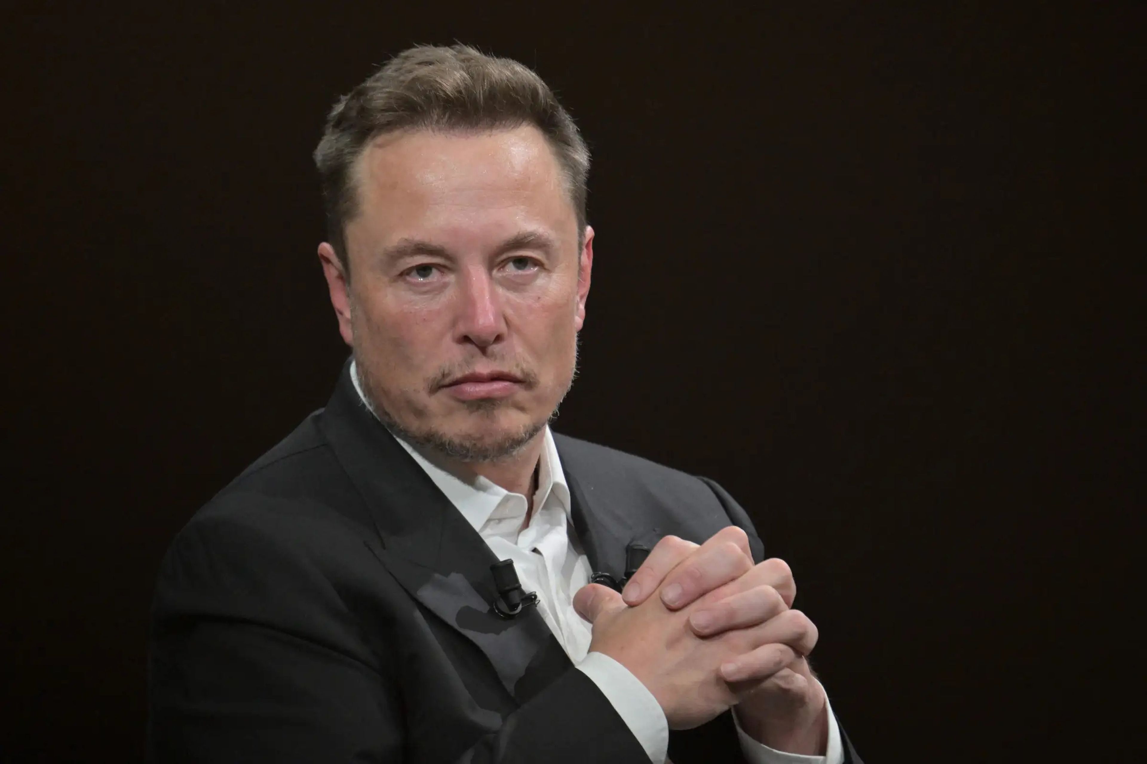 Elon Musk clasping his hands together with a serious expression on his face while wearing a black suit with a white button-down shirt.