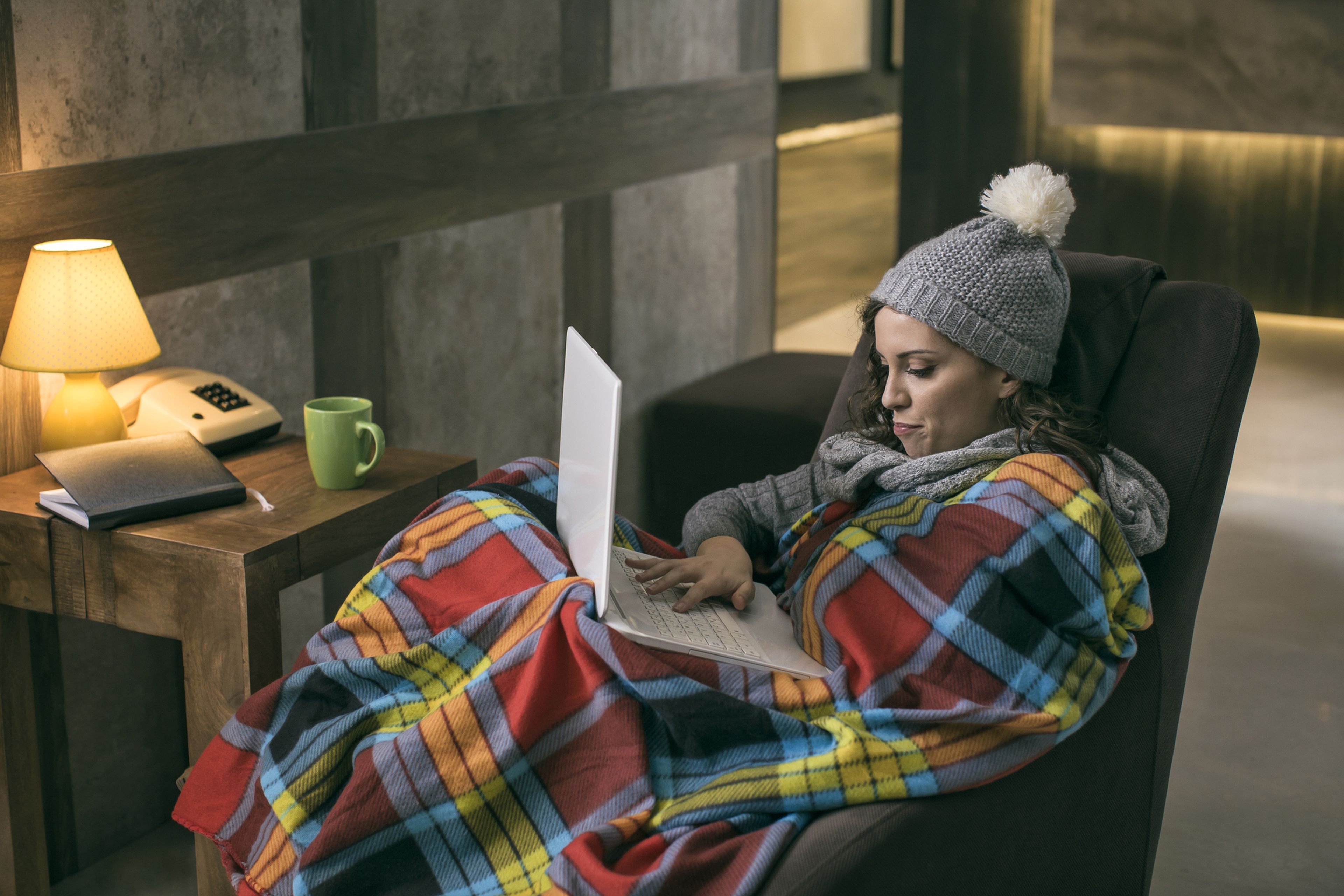 Una mujer trabaja en casa con el ordenador y tapada con una manta y un gorro.