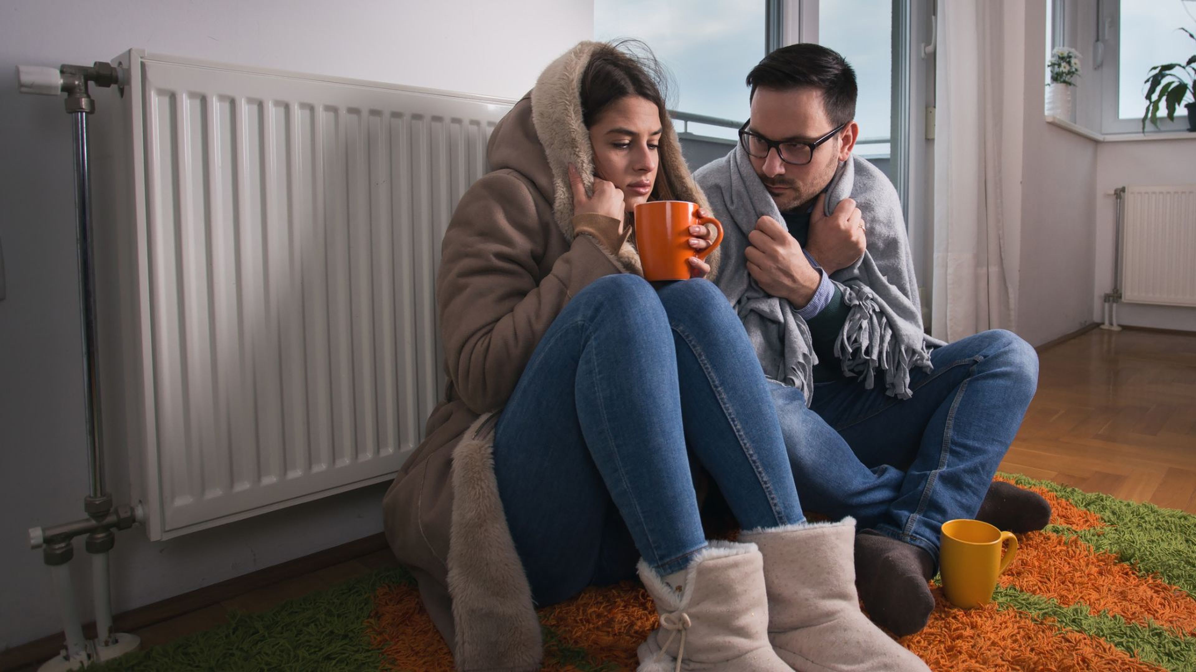 Una pareja sentada delante del radiador de su casa con mucho frío.