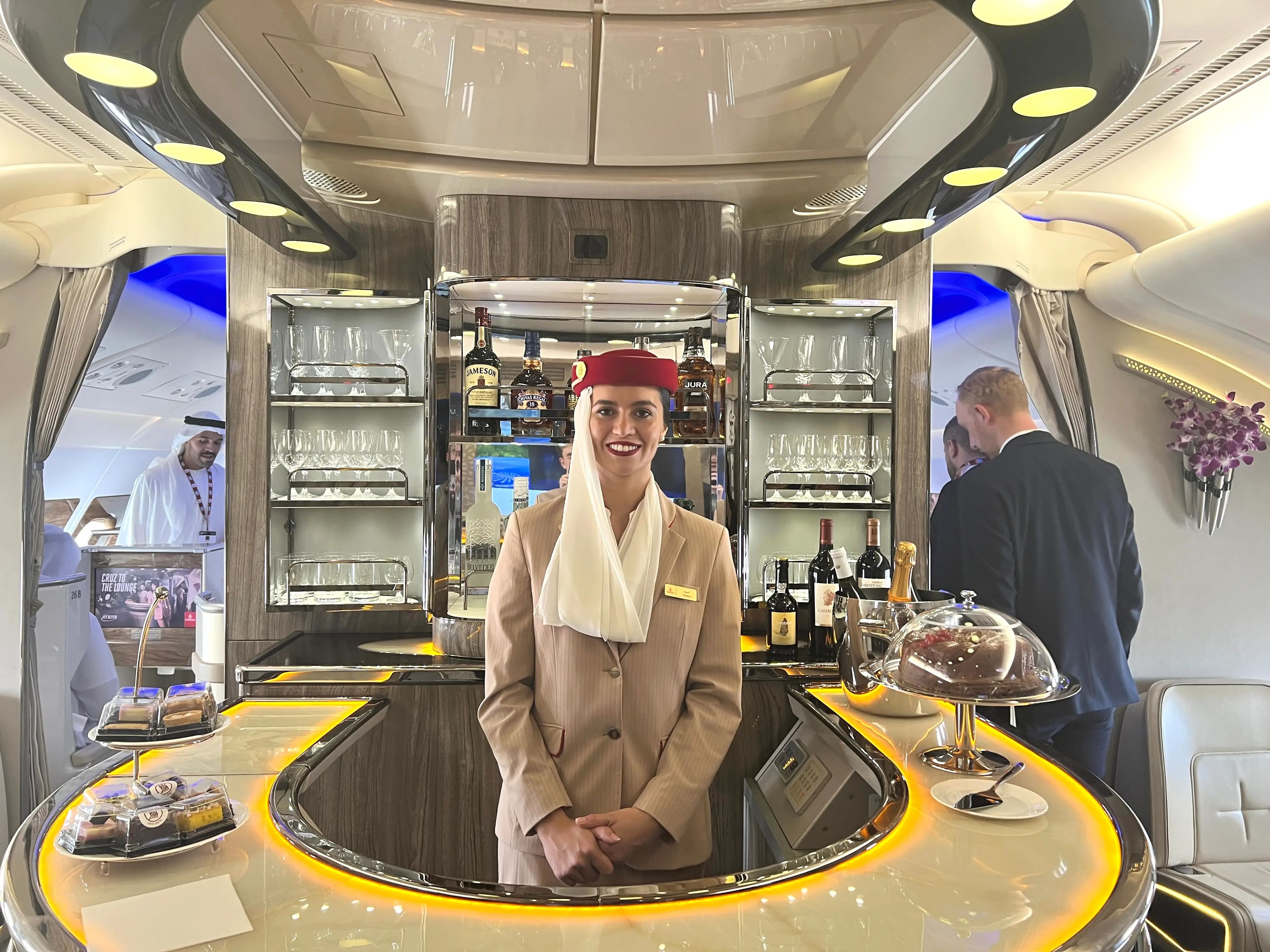 A Qatar Airways flight attendant smiling, stood behind the curved bar onboard an A380 with alcohol and glasses on the shelves and cakes on the bar.