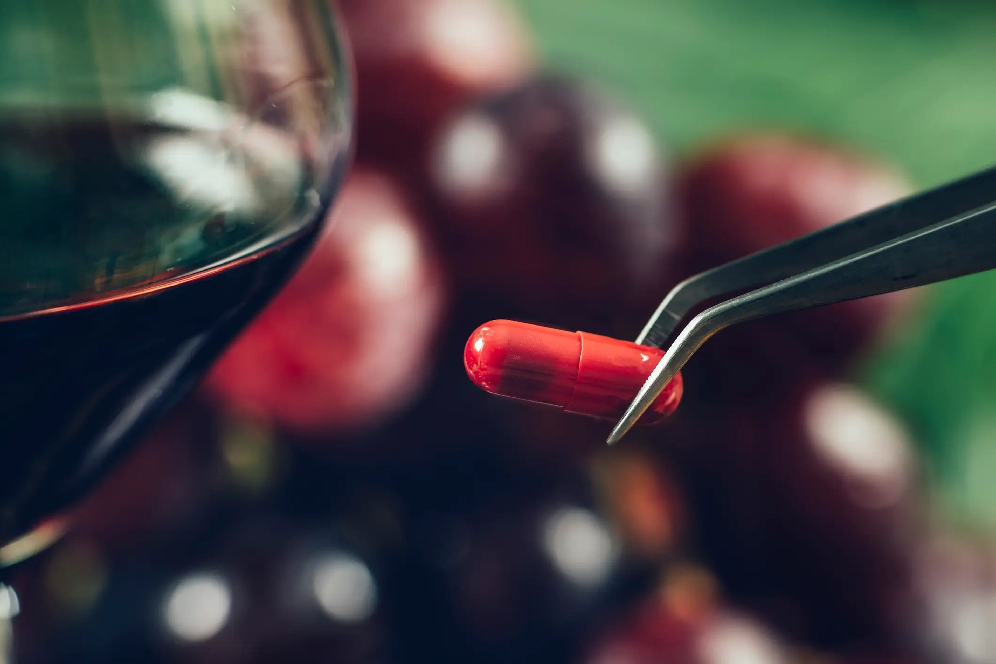 A red pill held with tweezers next to a glass of red wine.