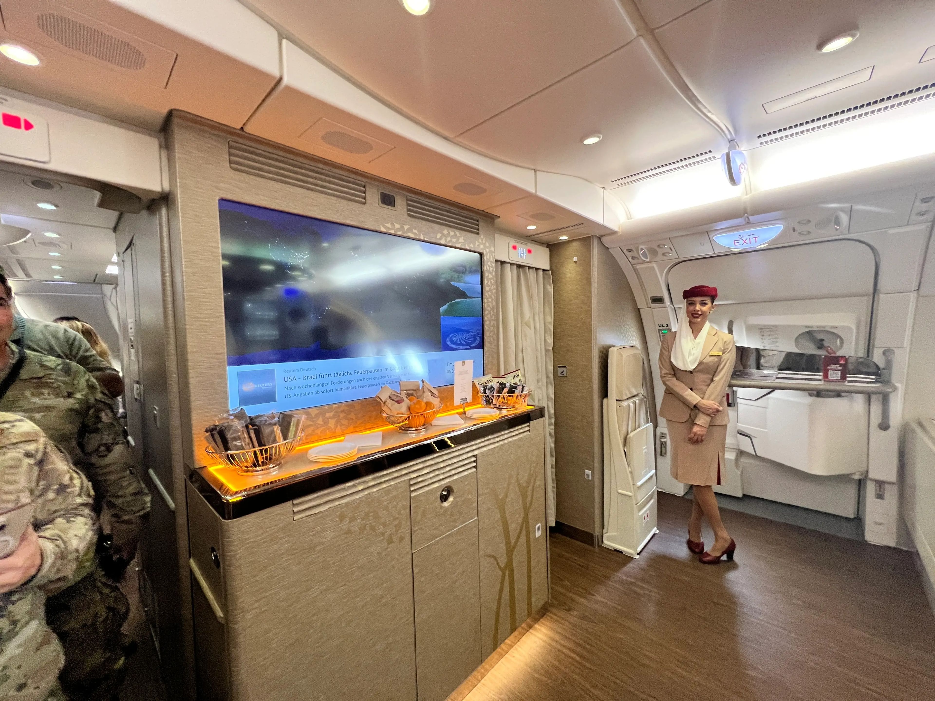 A reverse view of the bar area in an Emirates A380 shows a TV screen above a cabinet, and a flight attendant standing by the emergency exit.