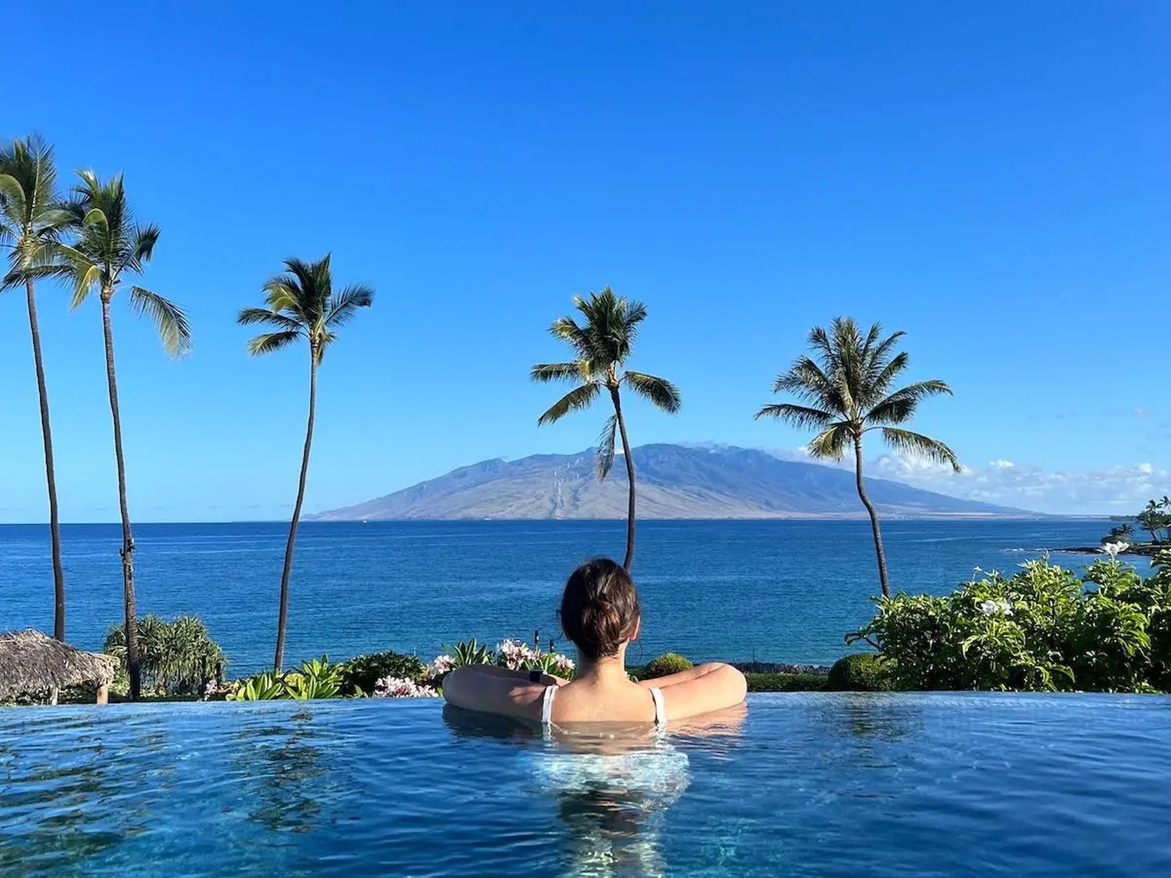 A woman with her back to the camera is pictured resting on the edge of an infinity pool, looking out at the Pacific Ocean and West Maui Mountains.