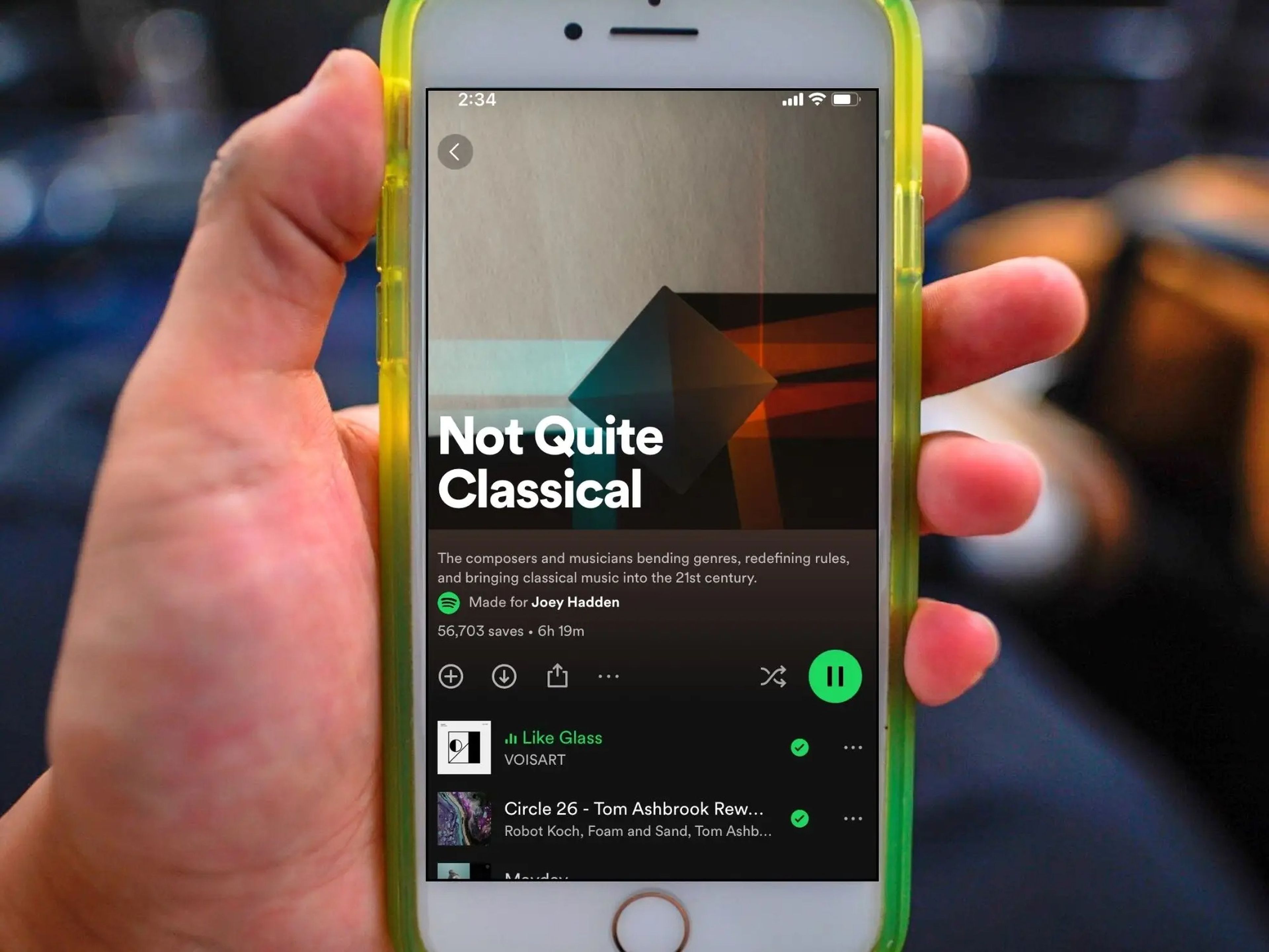A hand holds a white iPhone in a yellow case with a screen displaying a Spotify playlist called Not Quite Classical