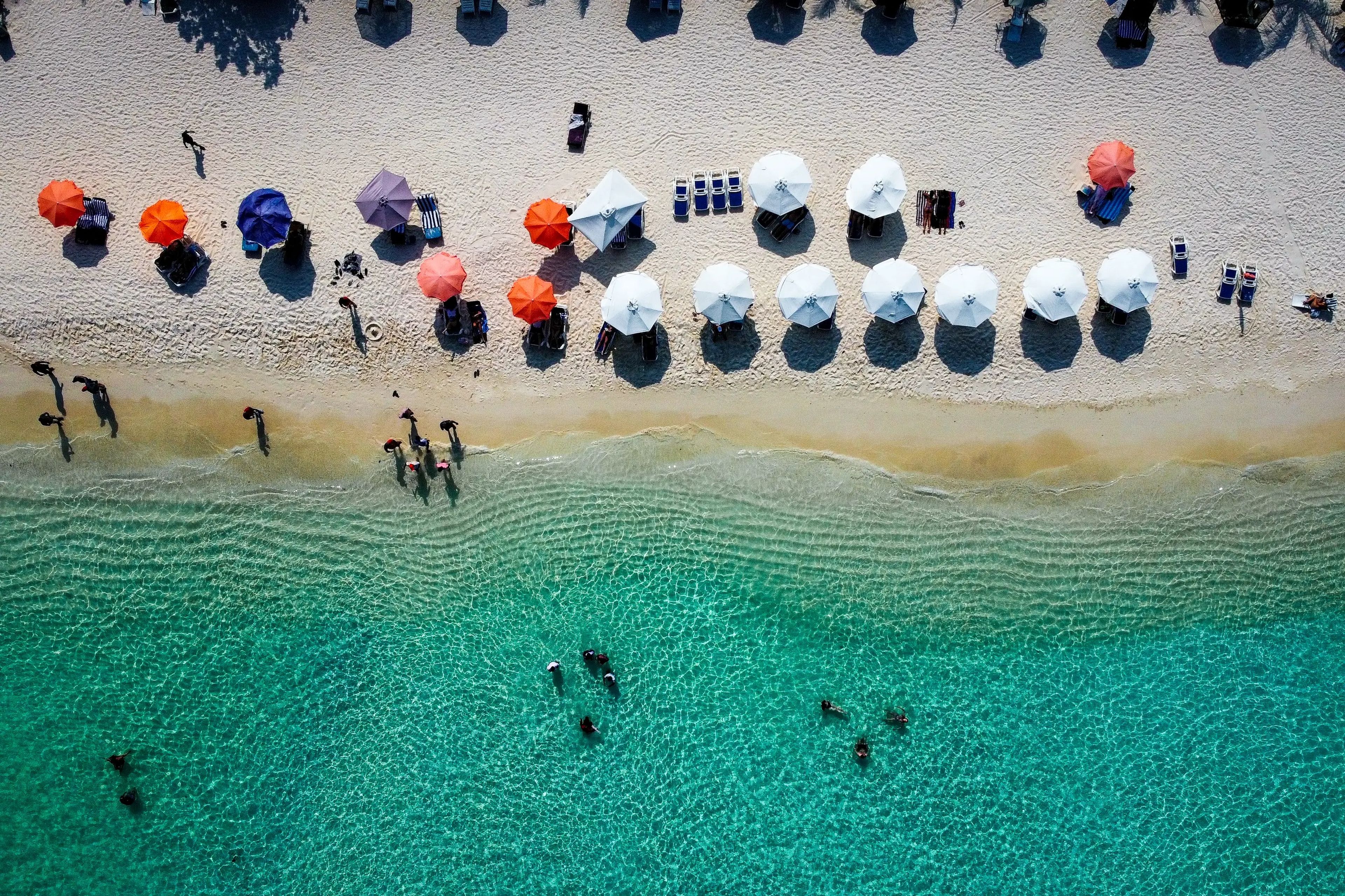 An aerial shot of Gulhi Island, with aqua-blue water and clean sand. There are multiple umbrellas on the beach, and people in the water and on the sand.