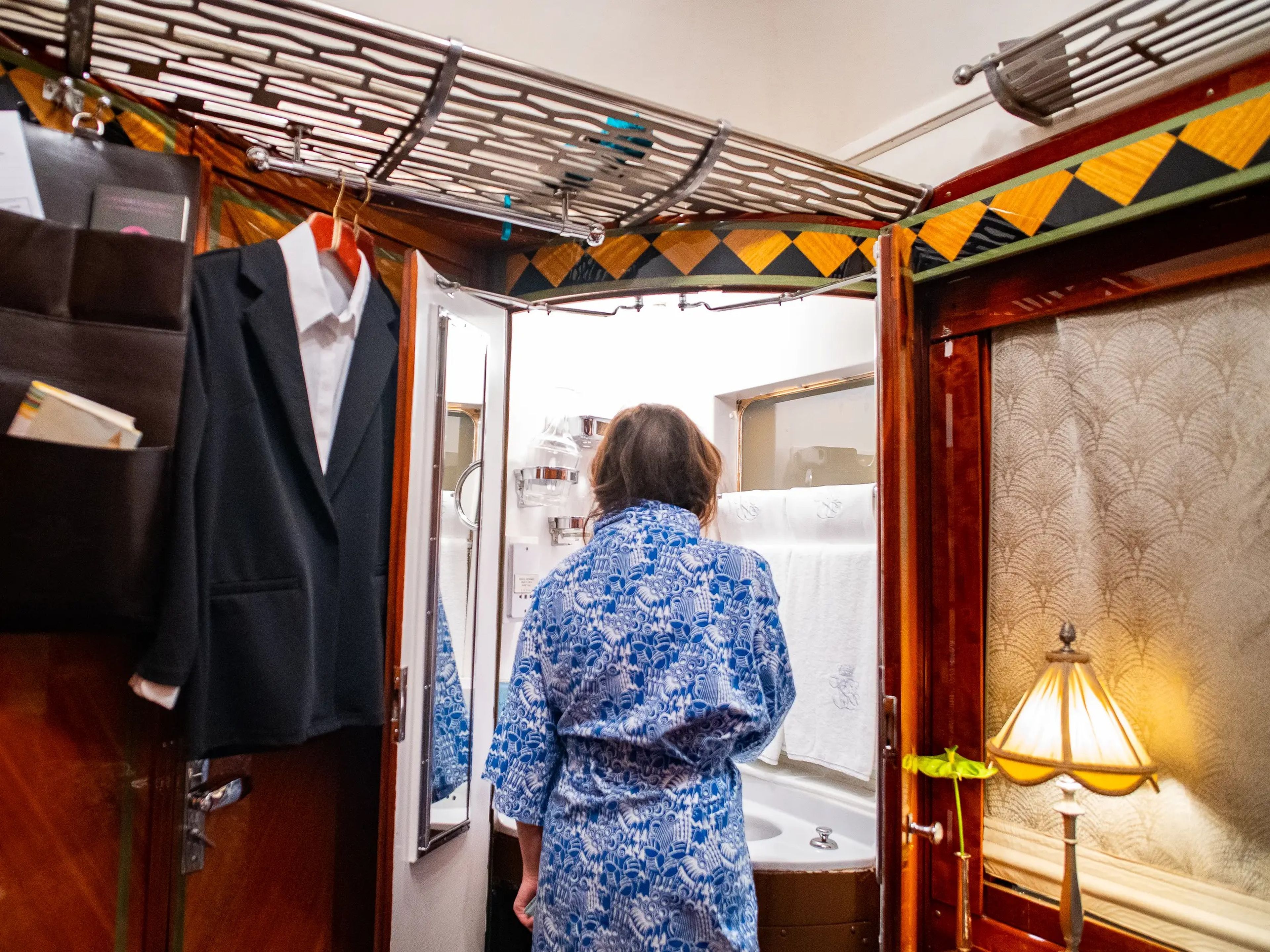 The author stands in front of a vanity of her train cabin with wooden finishes in a blue robe. There's a suit hanging on the left and a lamp on the right.
