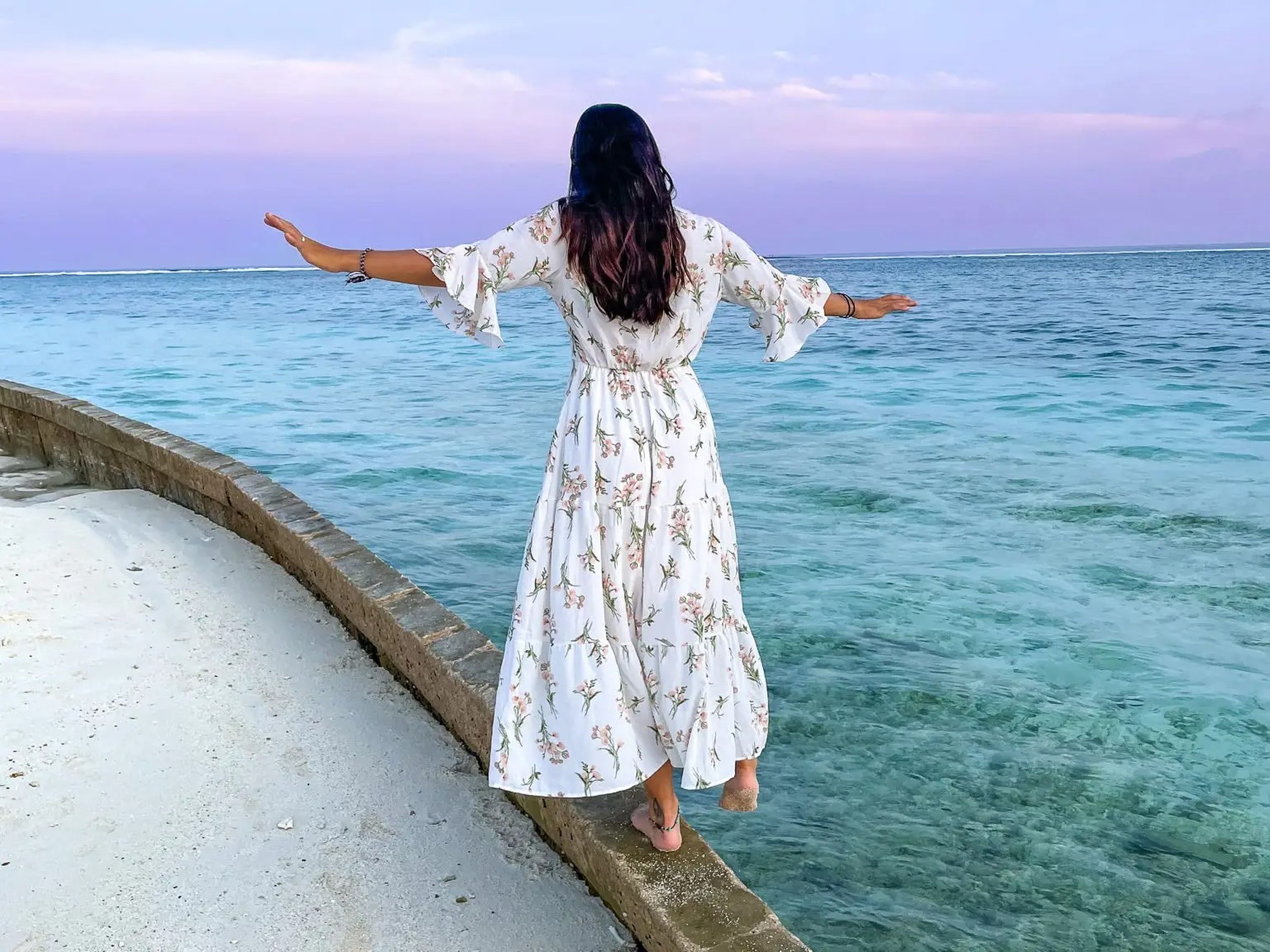 Geena balancing on a curb with her back toward the camera. Her foot is hanging over the turquoise water and the sky is different shades of purple