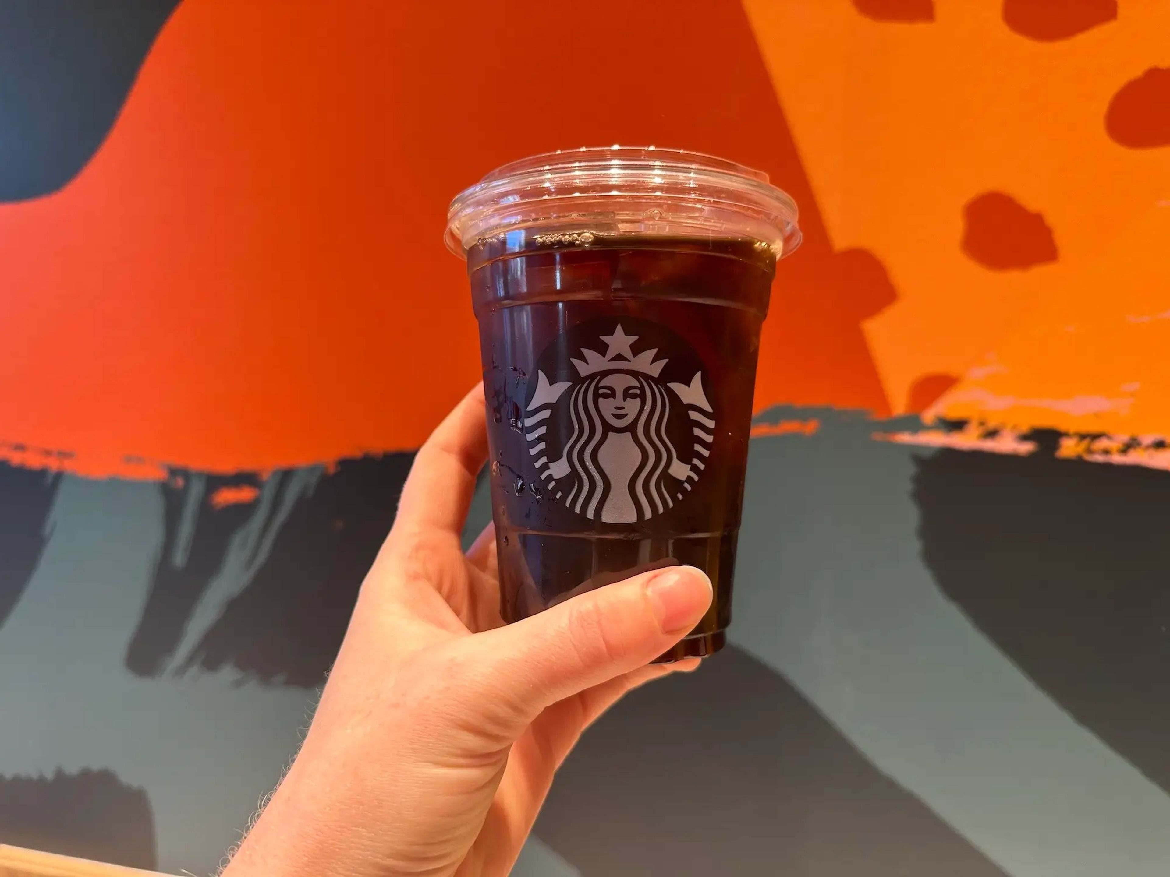 hand holding starbucks plastic cup with black iced coffee against a blue and orange patterned wall