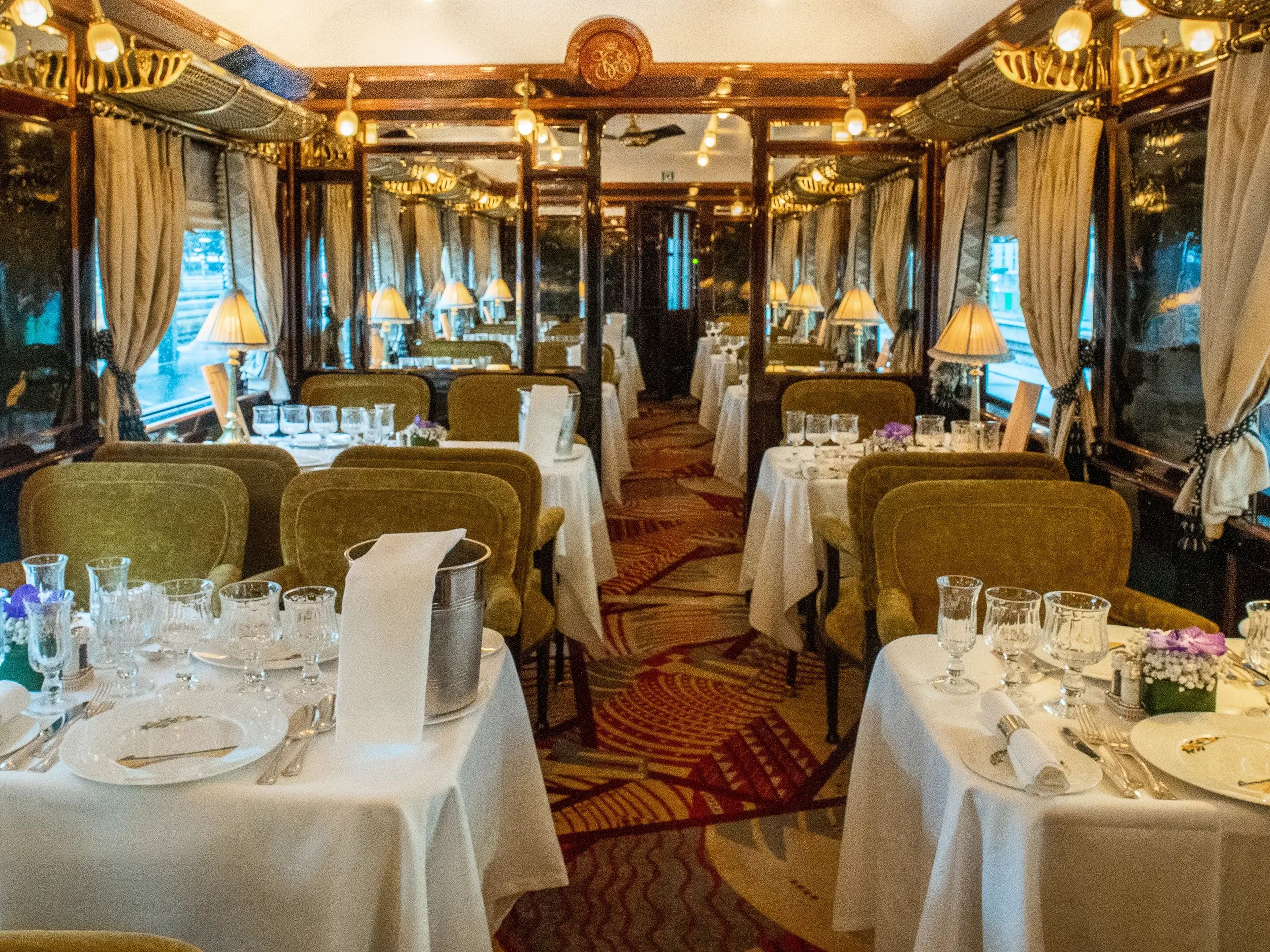 Inside a train dining car with velvet, beige seats, a warm-colored carpet, and tables with white cloths, dishes, and silverware