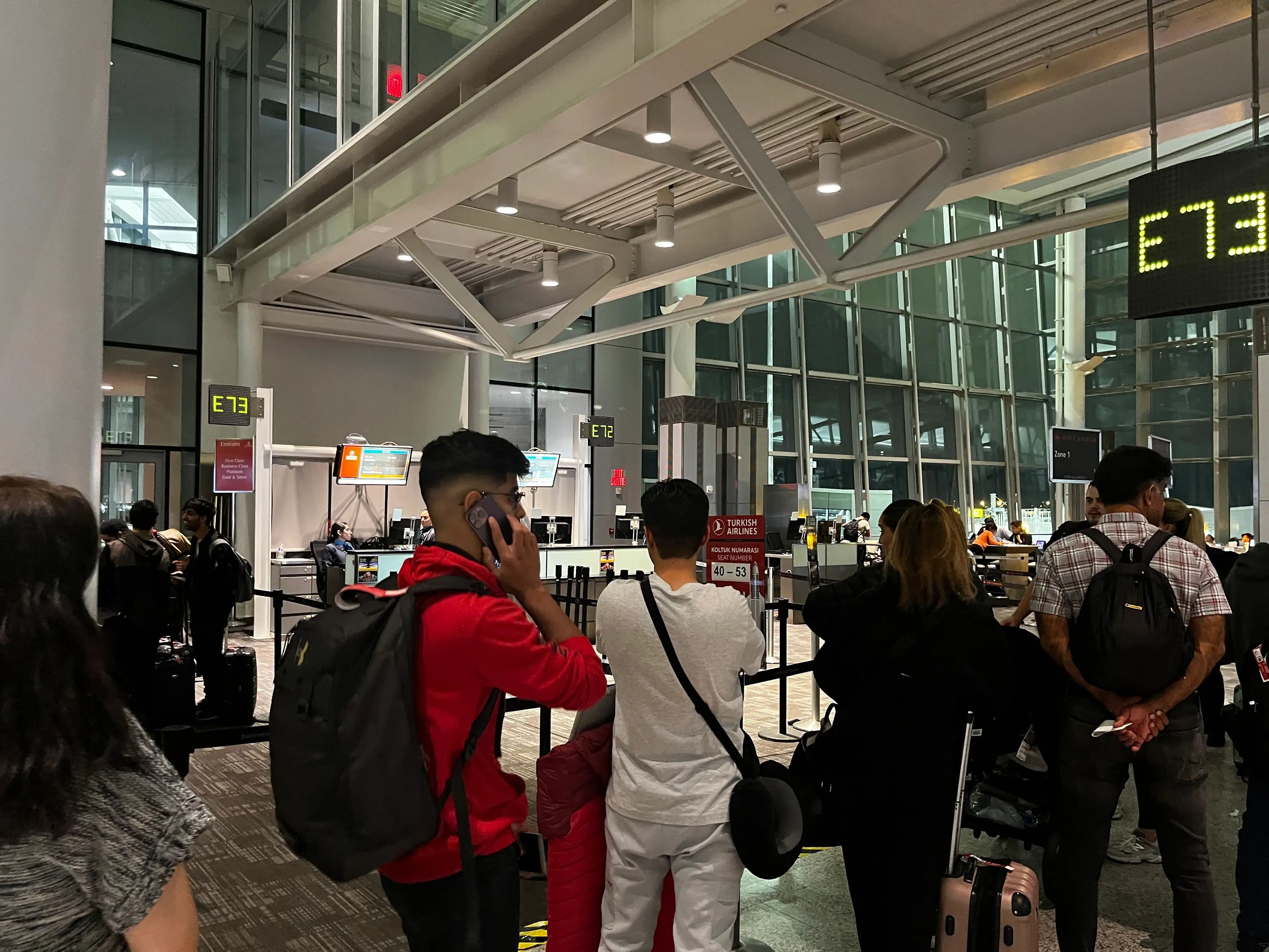 A line of people waiting to board in an airport with a man wearing a red shirt on the phone in foreground.