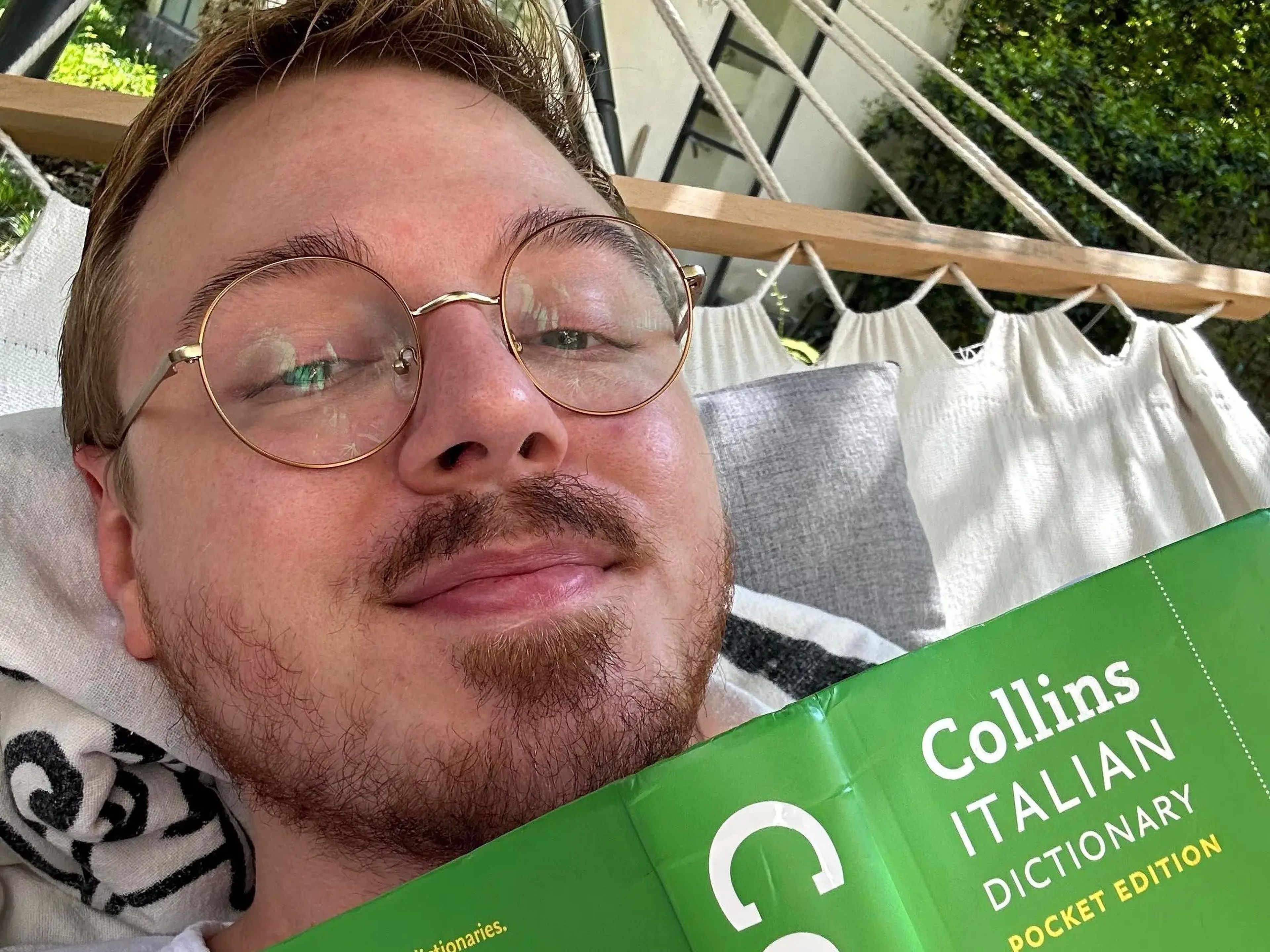 Man with glasses on a hammock holding a green Italian dictionary.