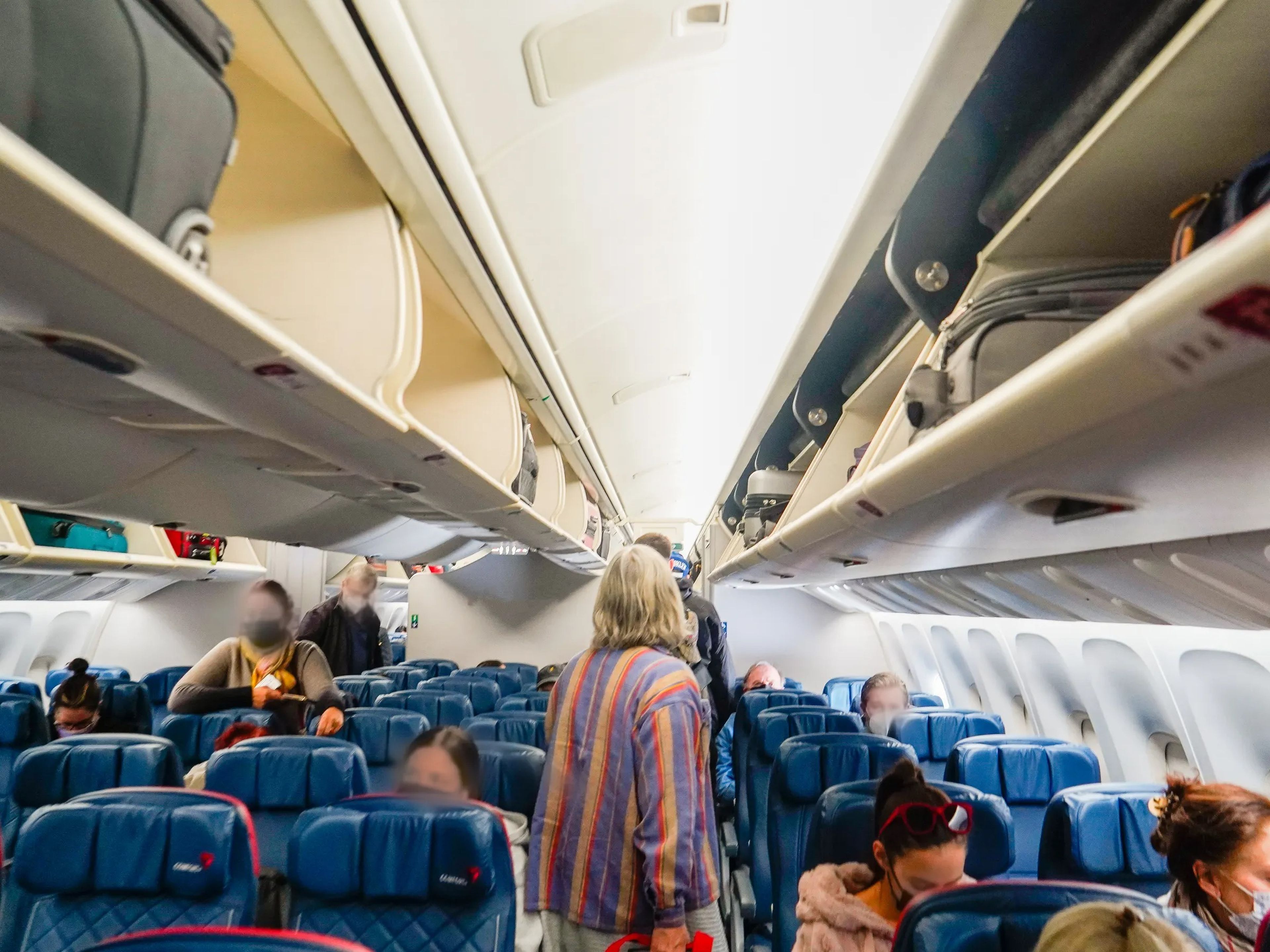 People board a plane with blue seats, luggages in overhead bin space are visible on the lop left and right