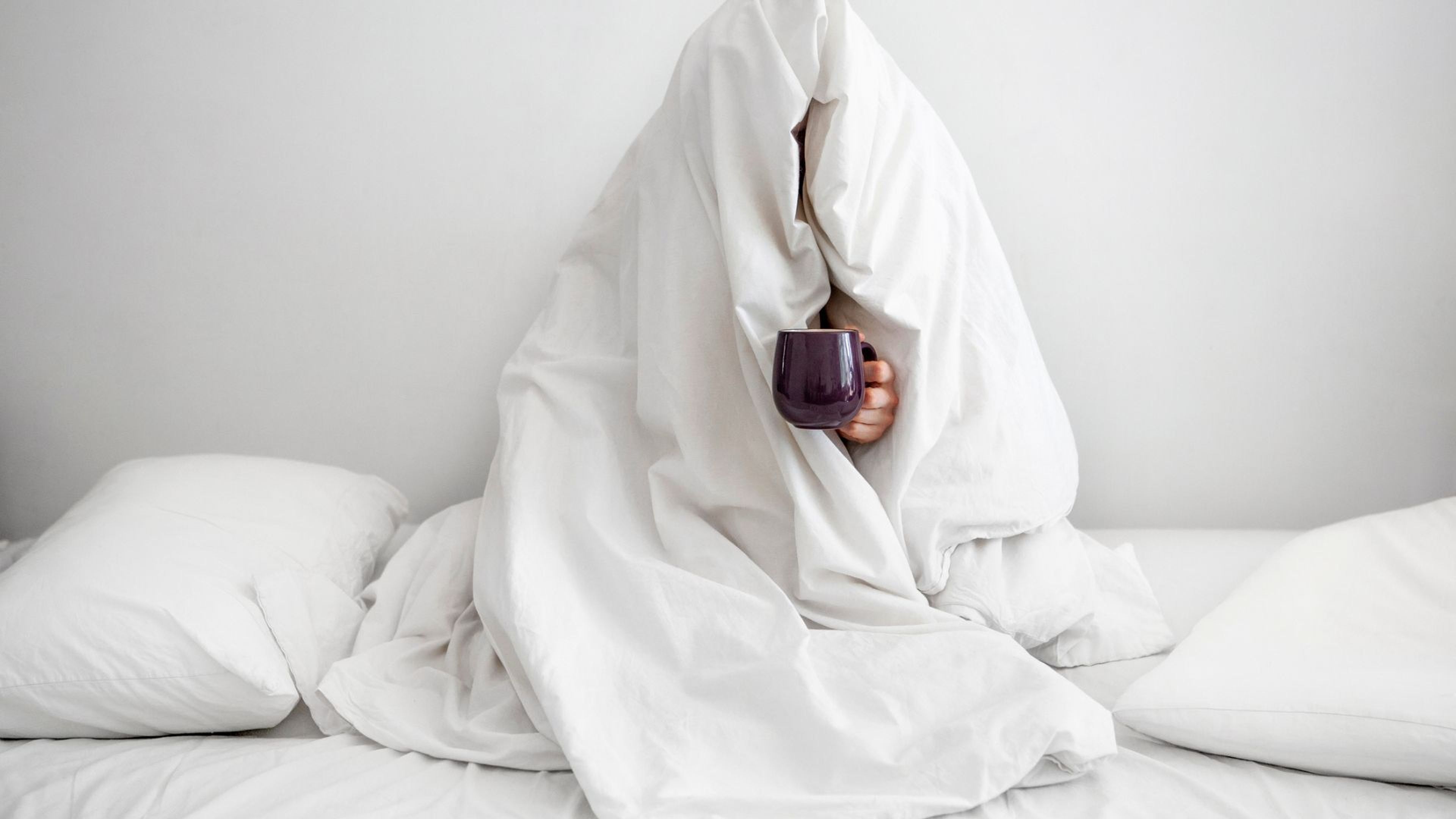 Una persona tapada de arriba a abajo, sentada en la cama y con una taza de café caliente.