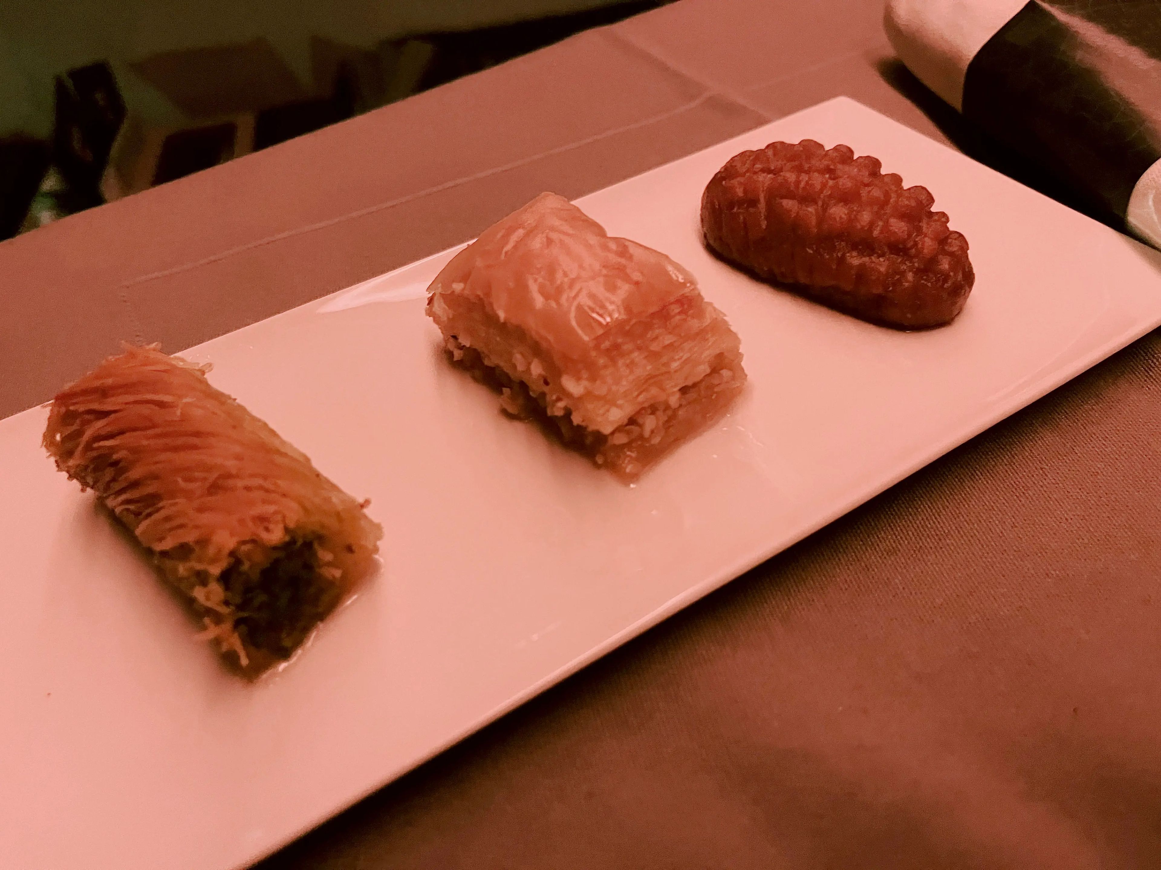 Pistachio kadayif (a cigar-rolled pastry), traditional baklava, and şekerpare (a fritter-shaped cake doused in syrup) lined up on a rectangular plate