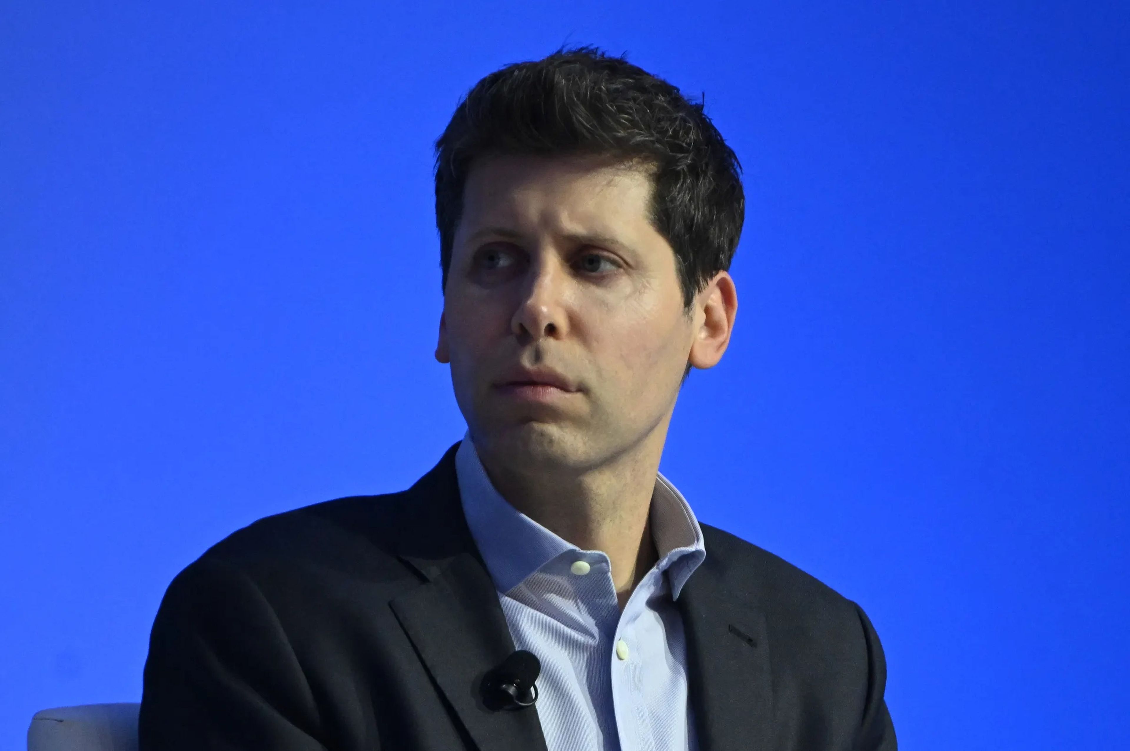 Sam Altman sits in front of a blue background, looking to the side.