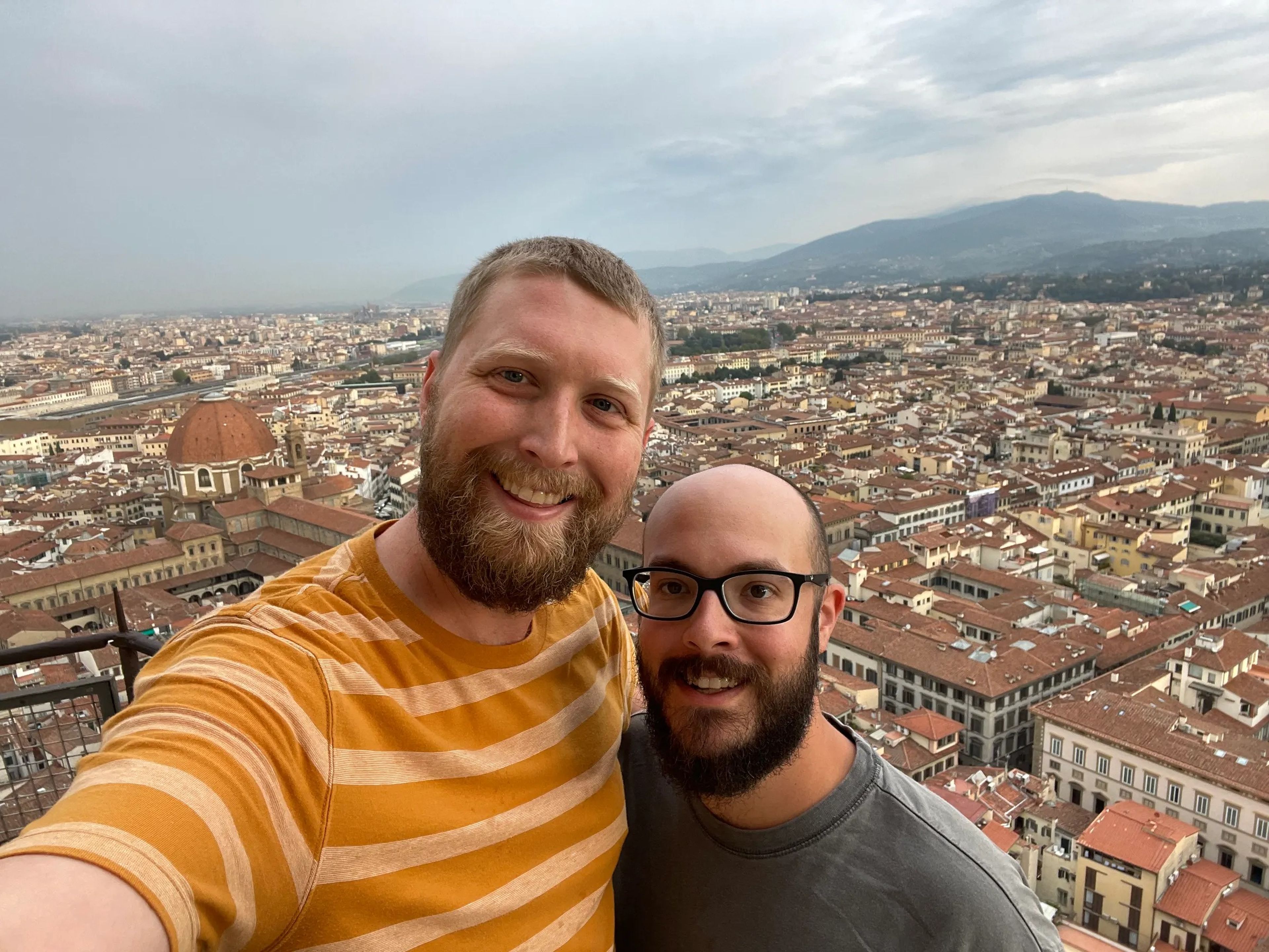 Selfie of the author wearing a yellow striped shirt and his husband in a gray shirt with houses in Florence, Italy, in background