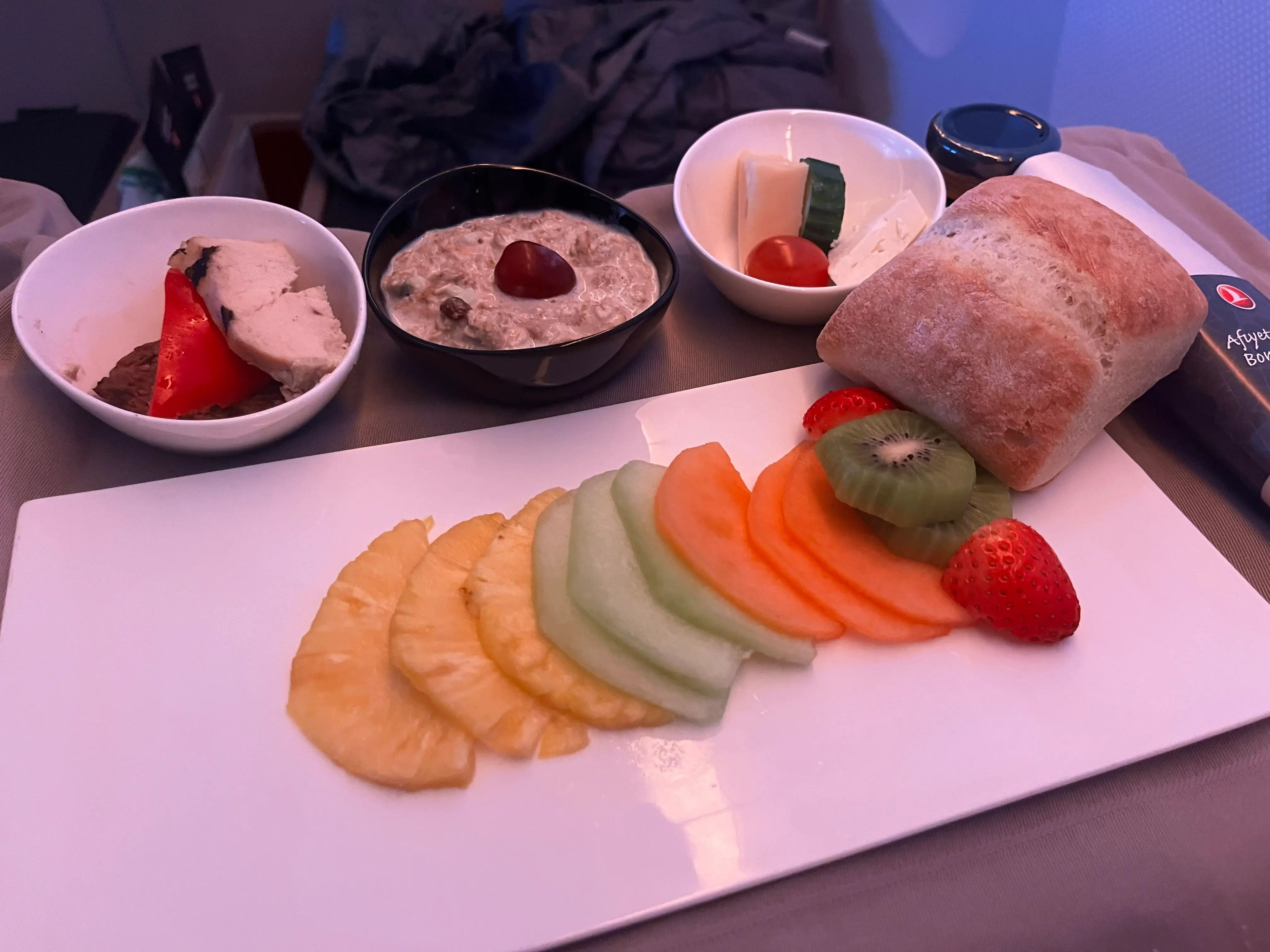 Spread of muesli, slices of kiwi, cantaloupe, melon, pineapple, and strawberry, chicken breast and roast beef, bread, and Turkish cheese with a tomato and cucumber