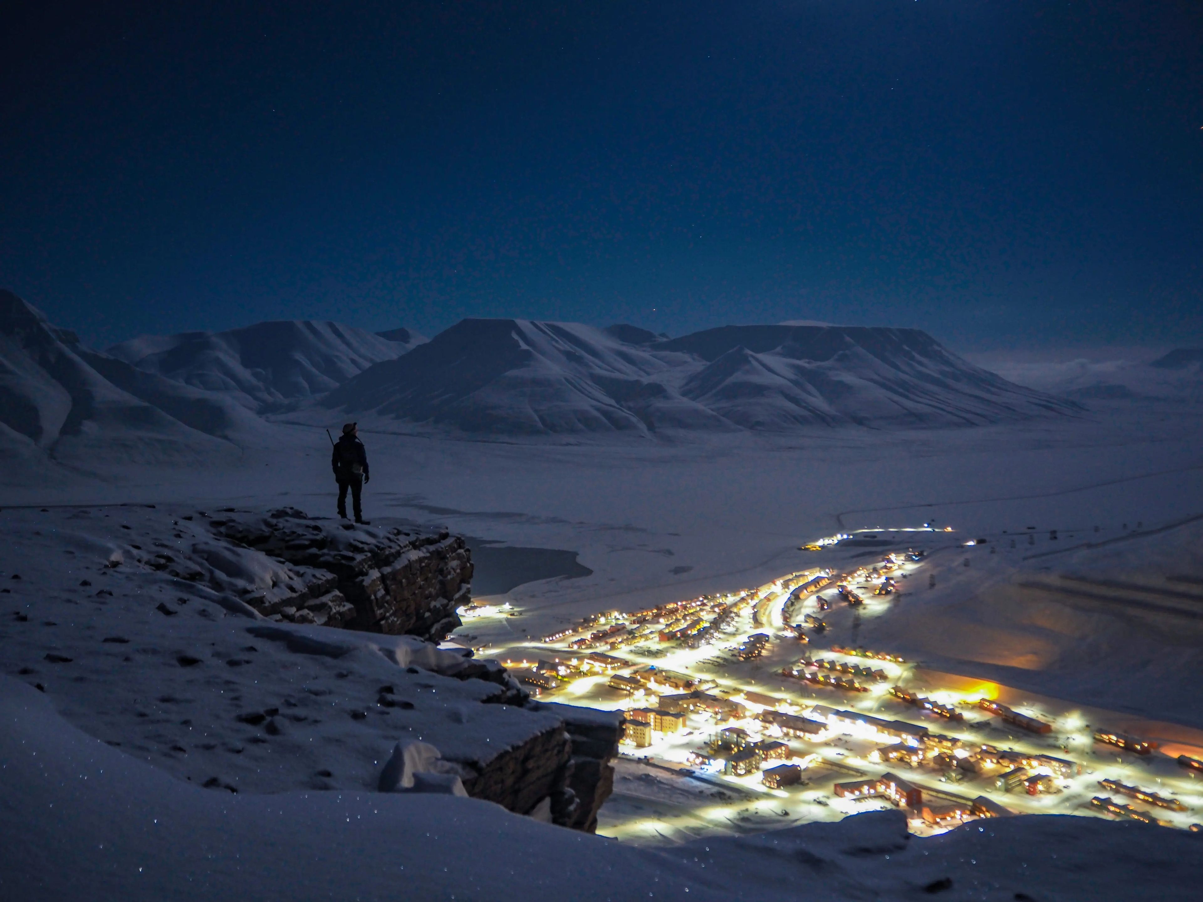 The writer stands on a mountain cliff overlooking a lit up town during the night
