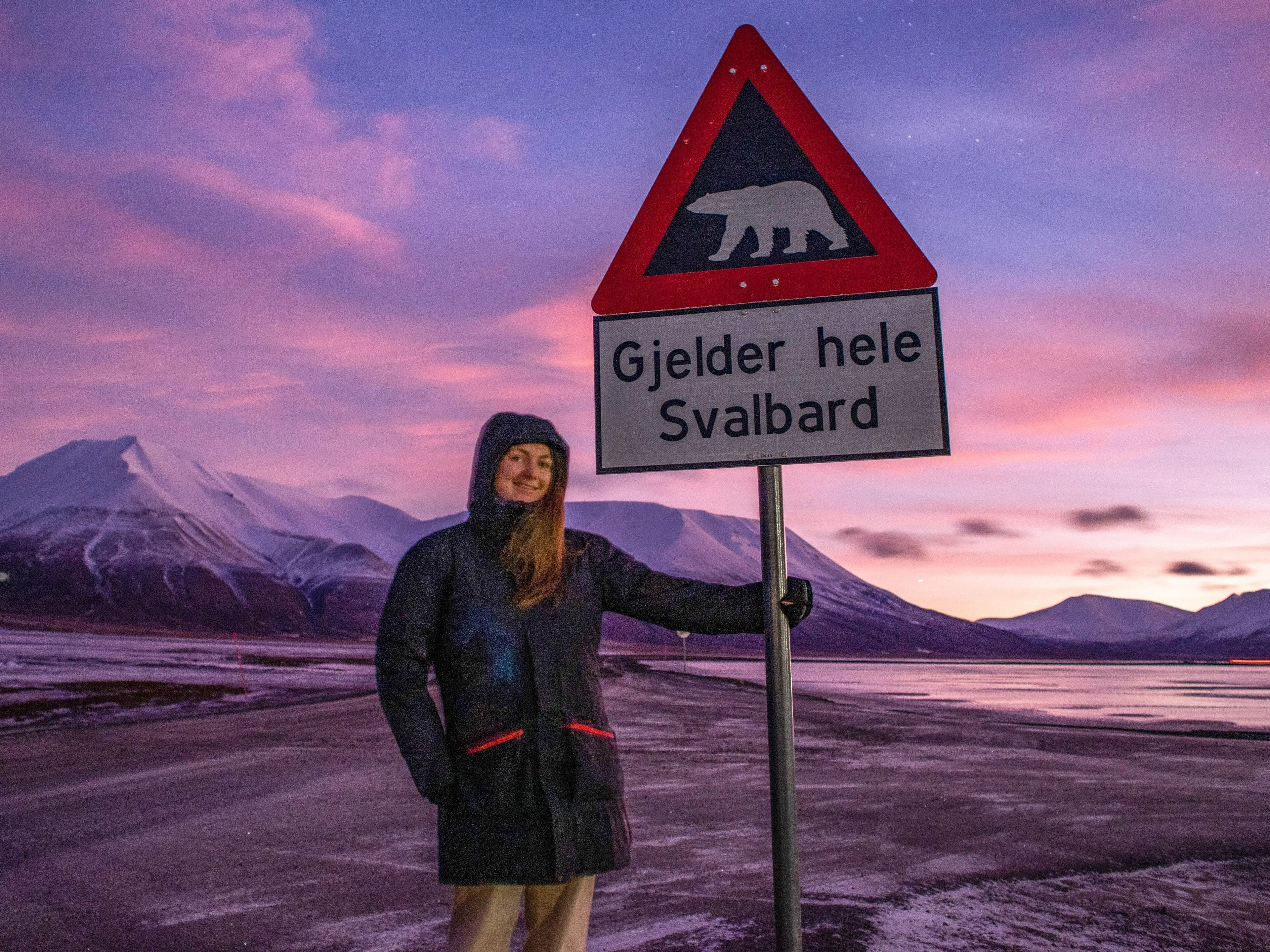 The writer stands next to a polar-bear zone sign with a pink sky in background