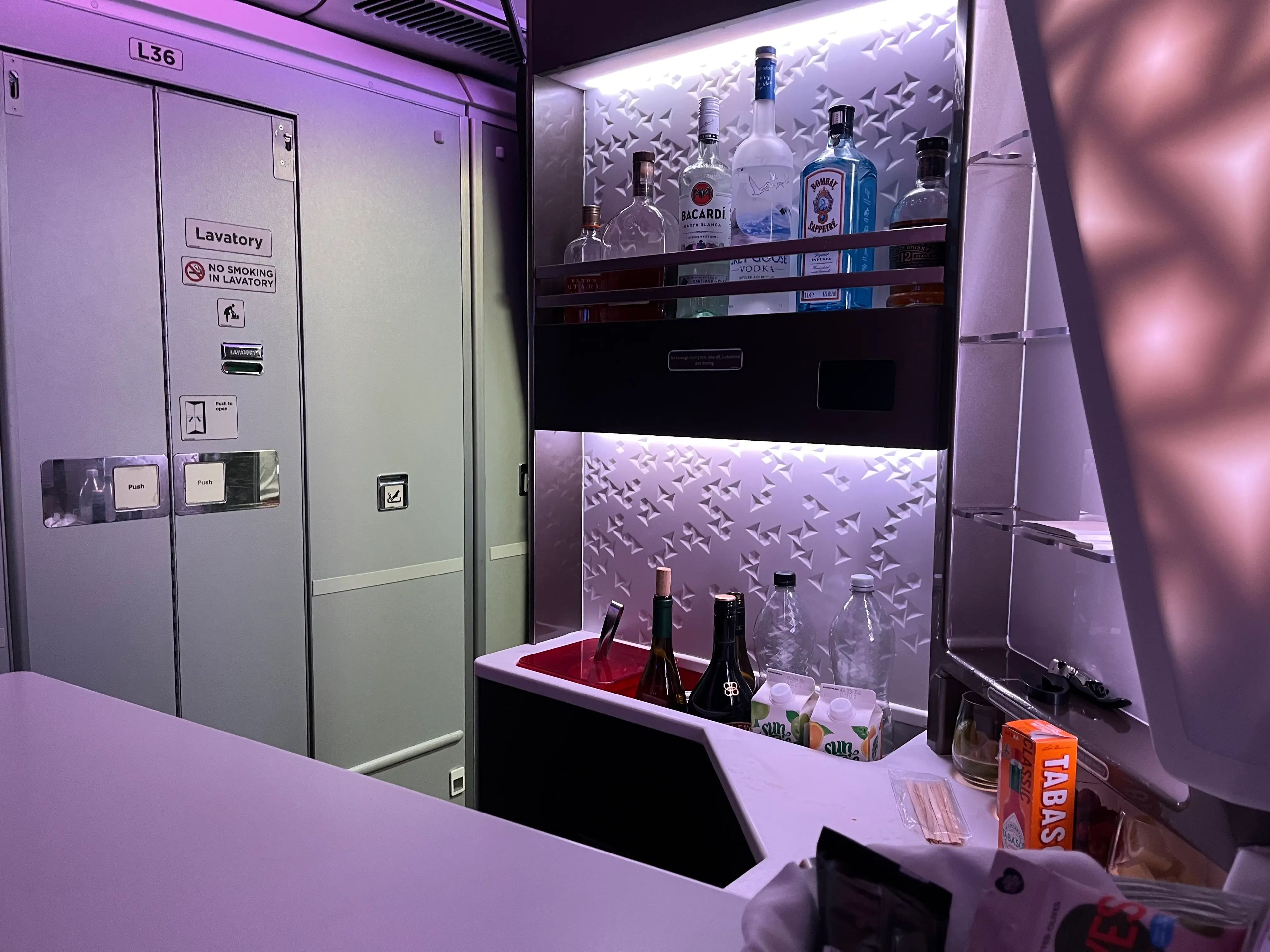 Airplane bar area with shelves stocked with Bacardi, Grey Goose, orange juice, and other mixers