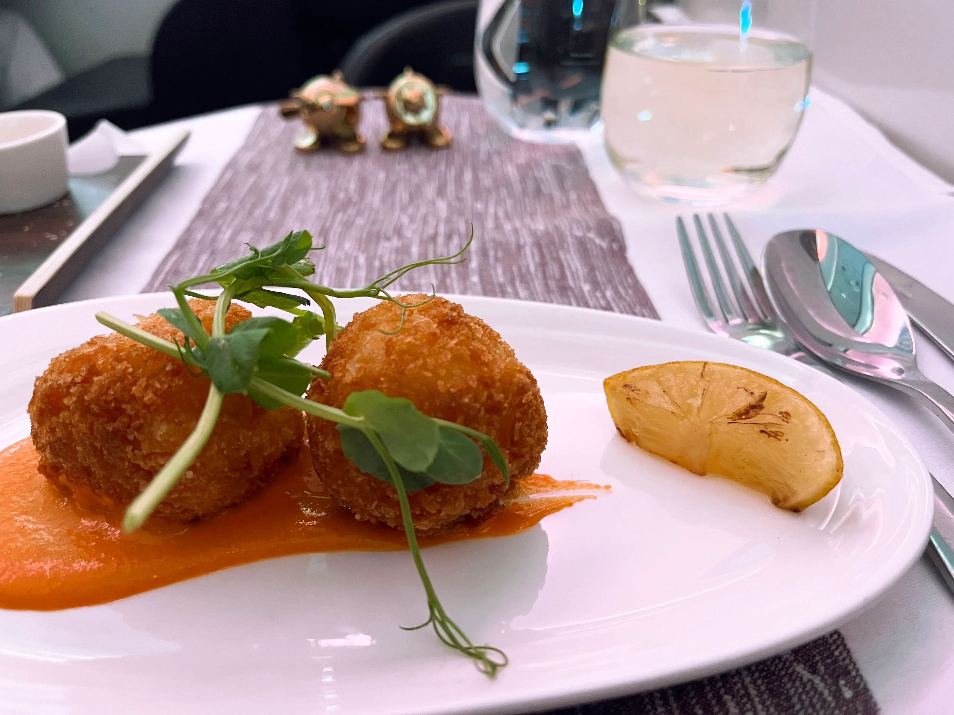 Butternut squash and sage arancini with sprigs of microgreens and a lemon wedge on the side. A table setting on an airplane tray is visible in the background