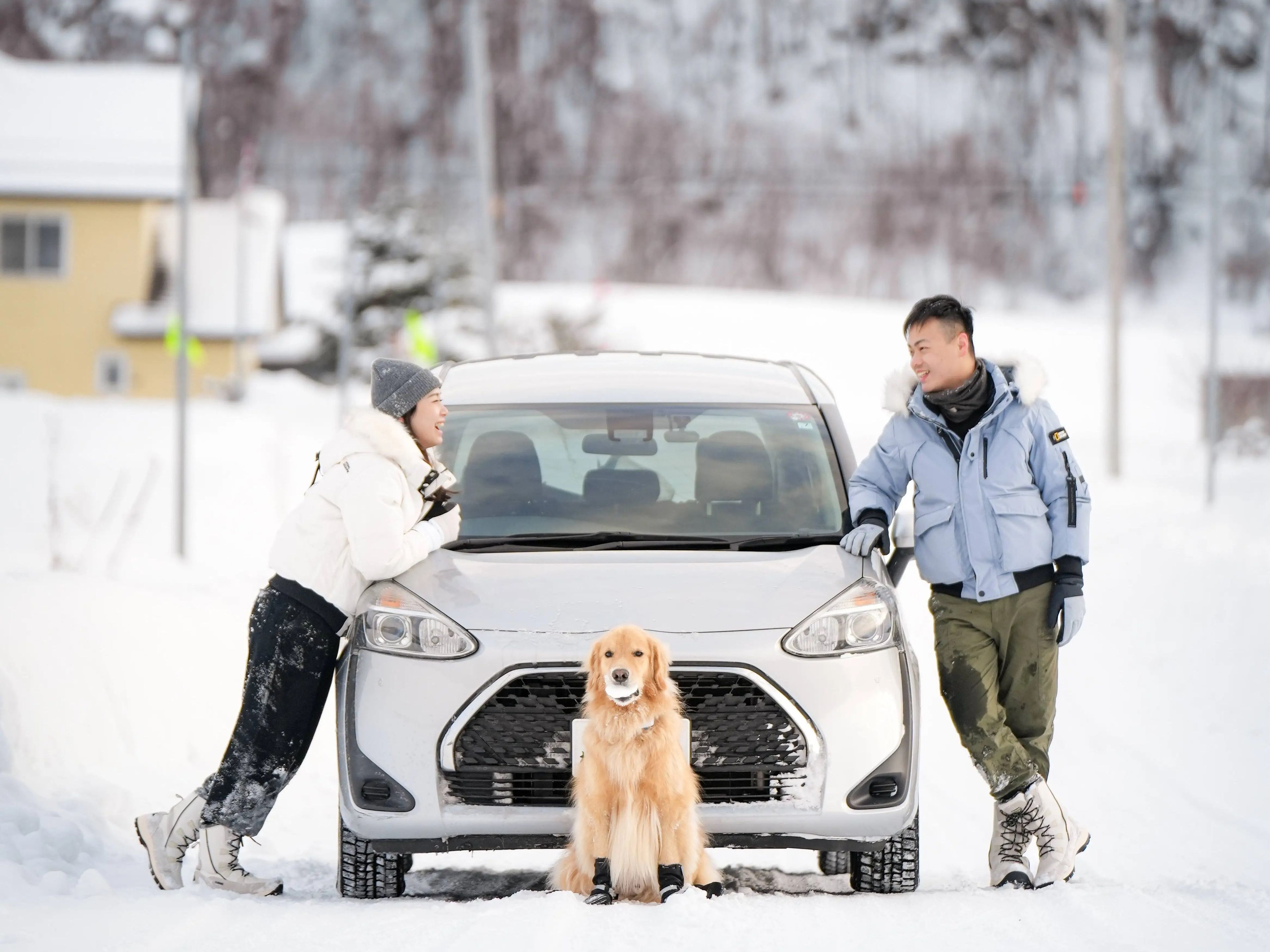Couple standing in the snow and leaning on a white car with Golden Retriever in front.