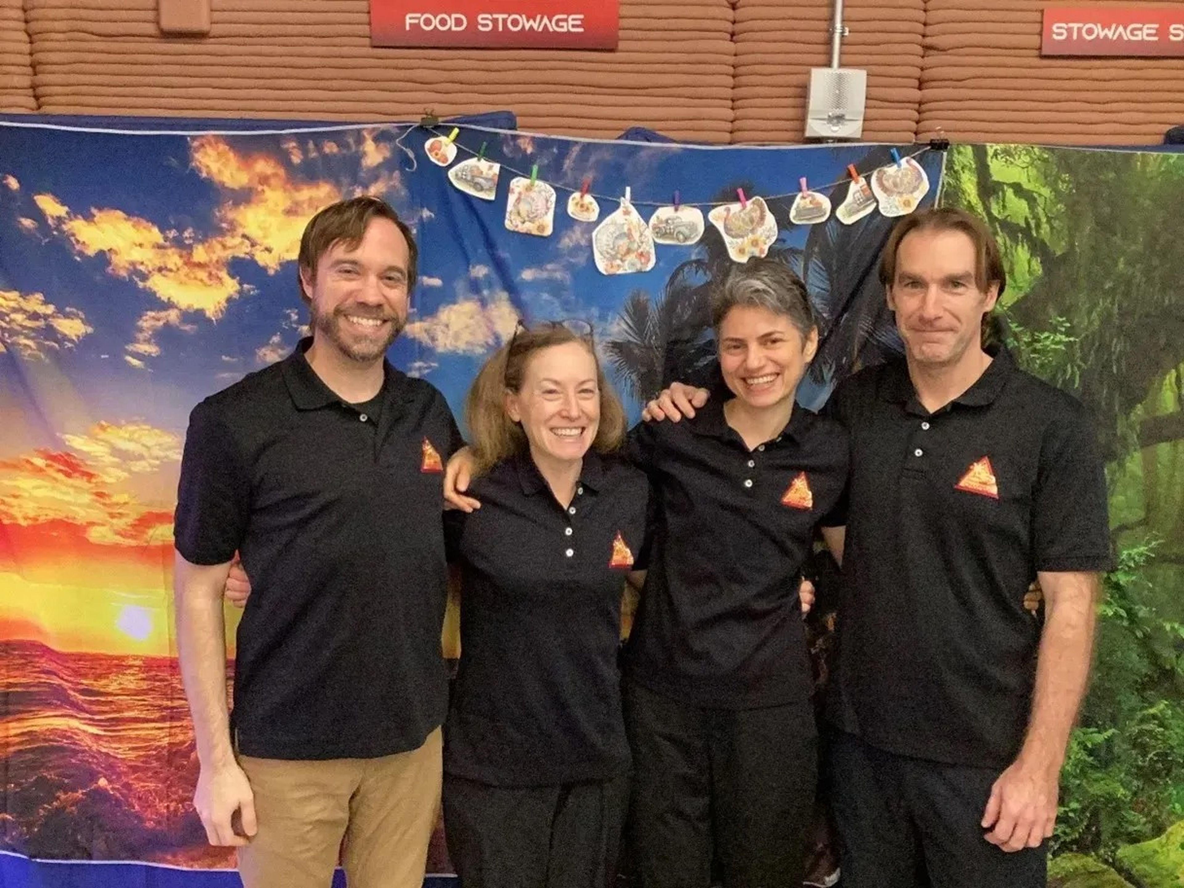 four people wearing black polo shirts with the same small orange triangle logo pose together in front of a hanging tapestry of an island sunset with thanksgiving turkey drawings pinned at the top