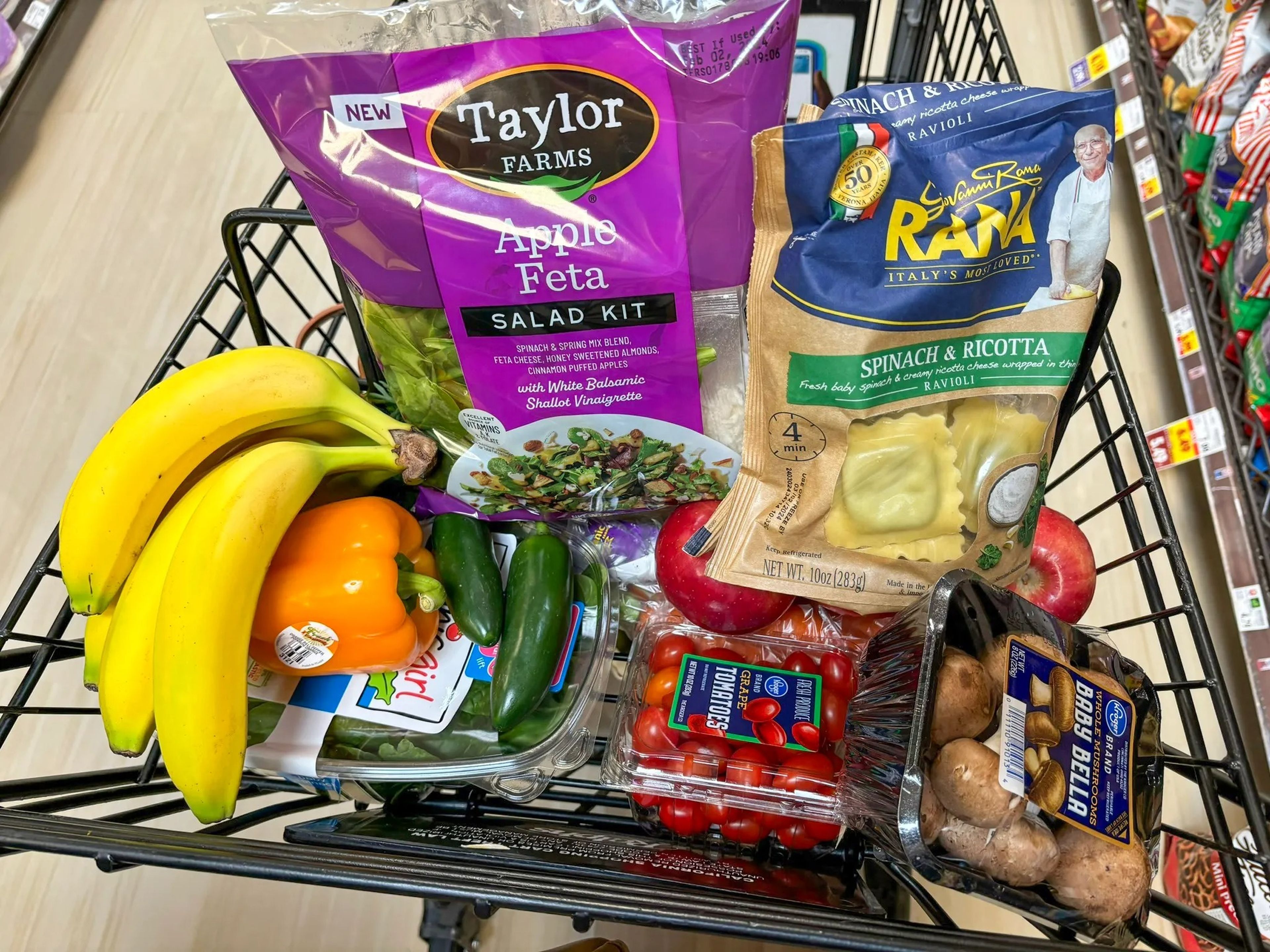 A grocery cart filled with tomatoes, apples, a package of mushrooms, peppers, bananas, an apple-feta salad kit with a purple label, and a bag of Rana spinach-and-ricotta pasta