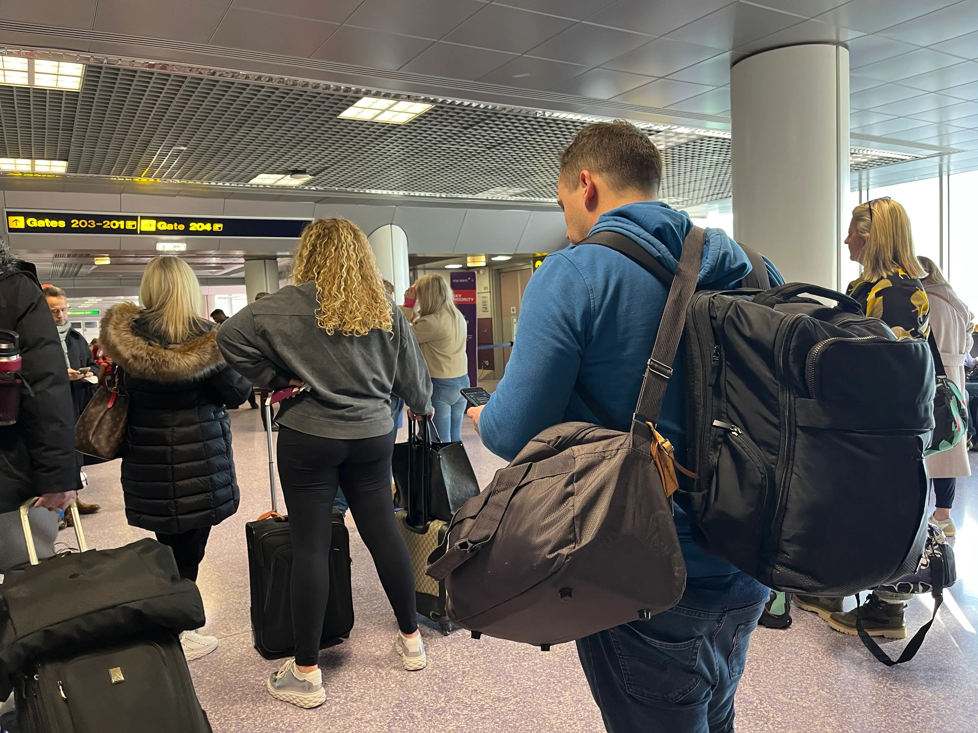 Line of people waiting to board Virgin Atlantic flight. Some passengers include a man in a blue hoodie, a woman with curly blonde hair in a gray sweatshirt, and a woman with a black puffer jacket