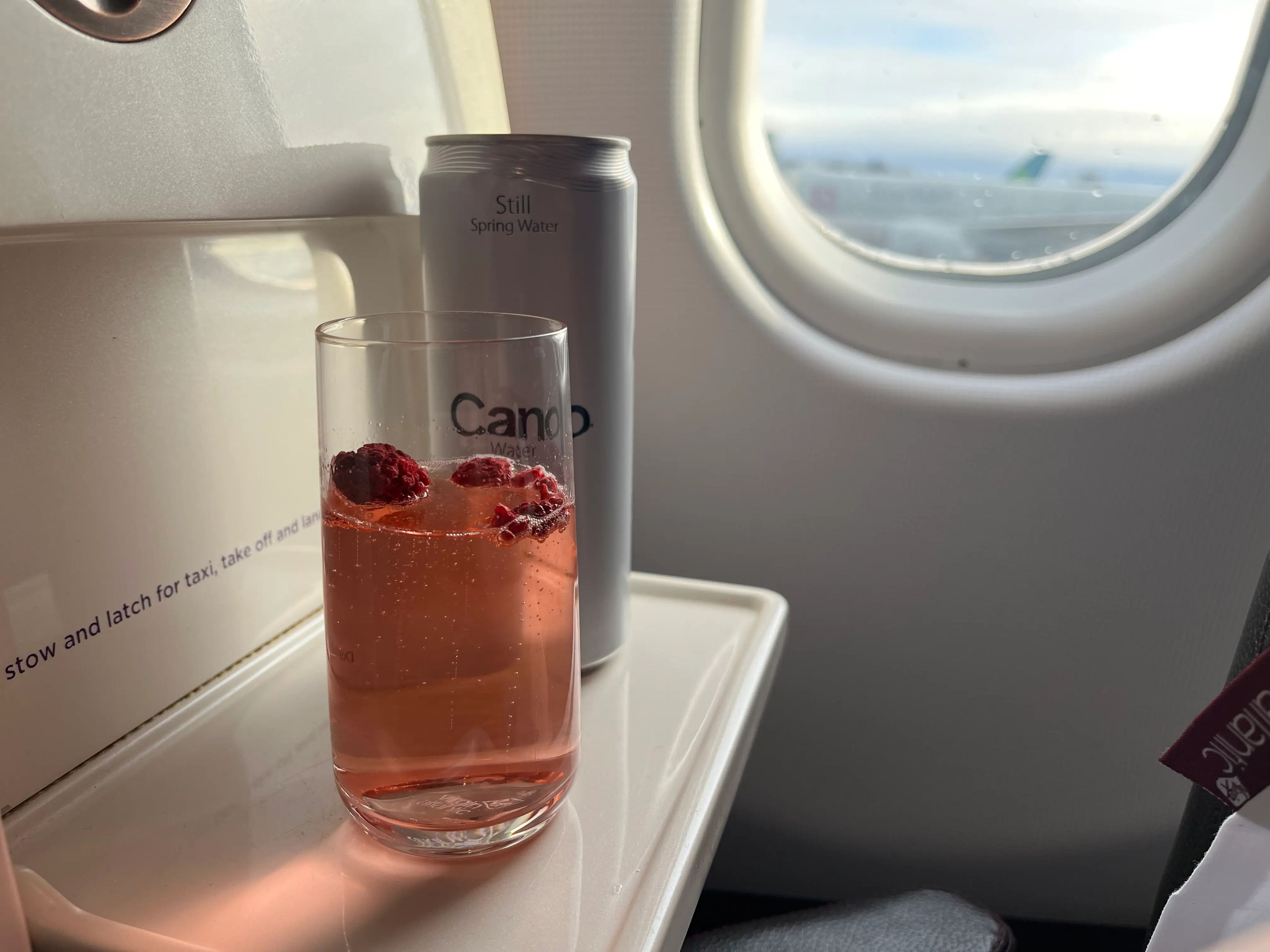 A red-orange cocktail with sparkling wine and raspberries floating at the top alongside a gray can of spring water on a tray on an airplane with window in background