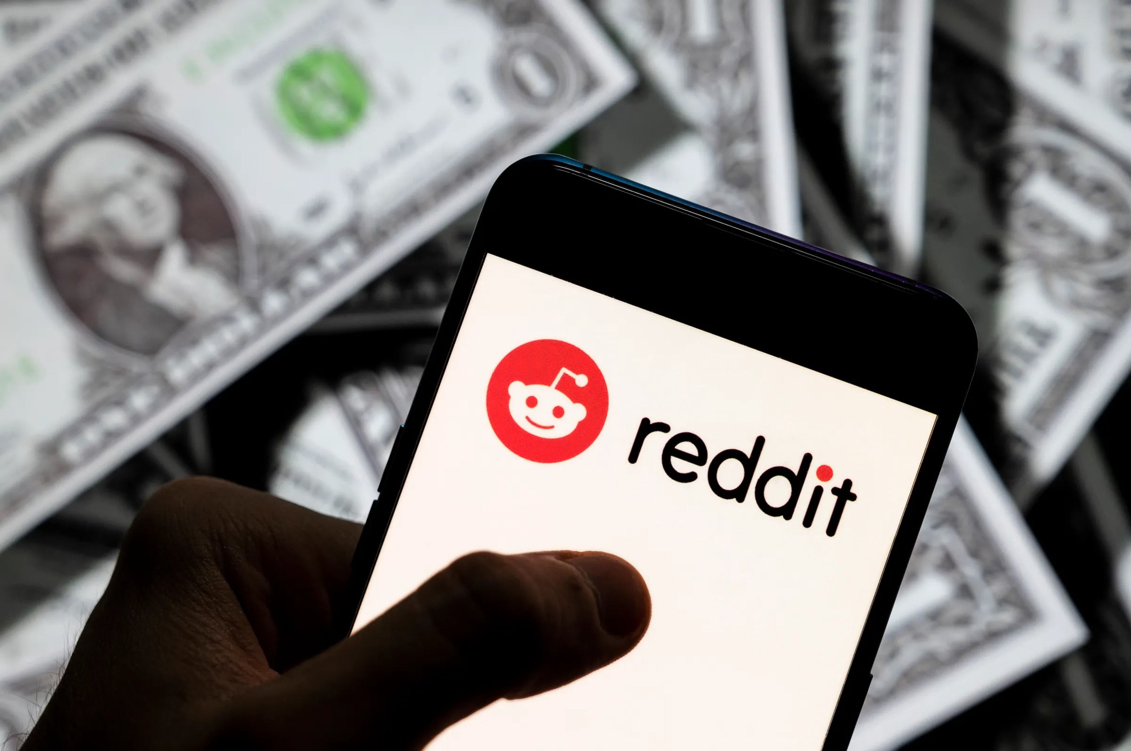 Reddit logo on a cell phone with images of dollar bill in the background.