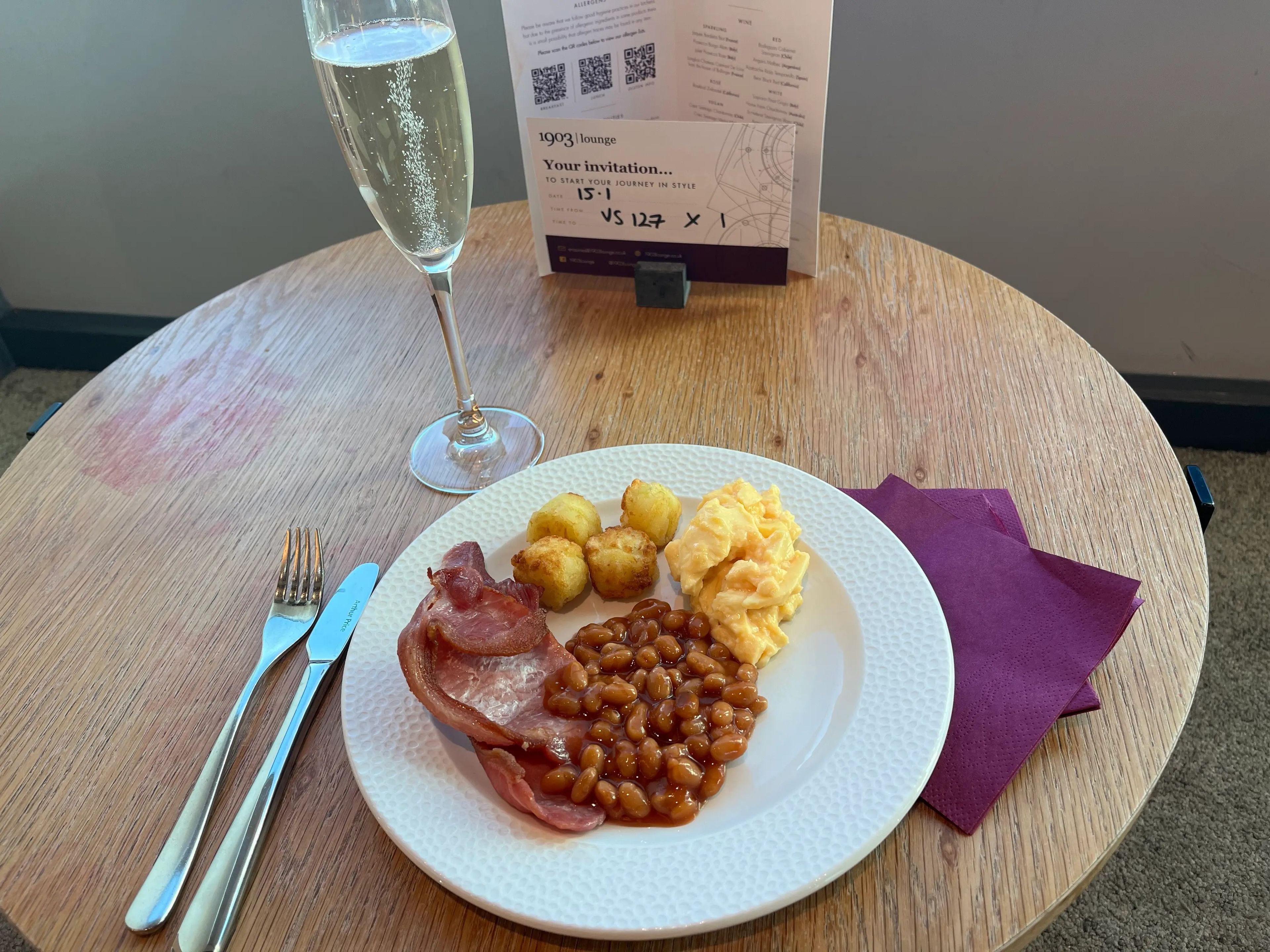 Round circular faux wooden table with a glass of sparking wine and a white plate of ham, baked beans, scrambled eggs, and tater tots