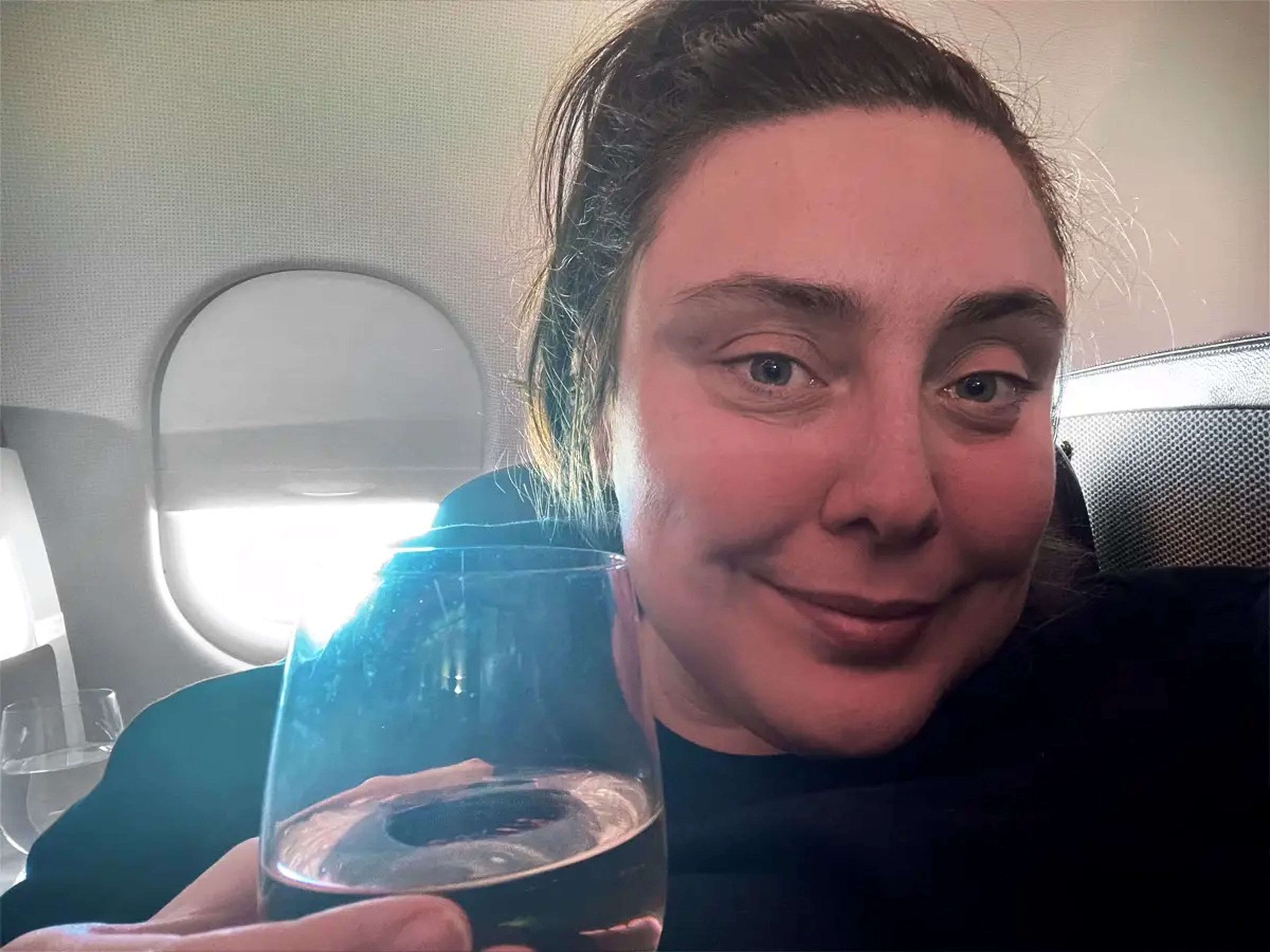 Selfie of the writer smiling, holding a glass of wine, and the airplane window with light shining through in the background