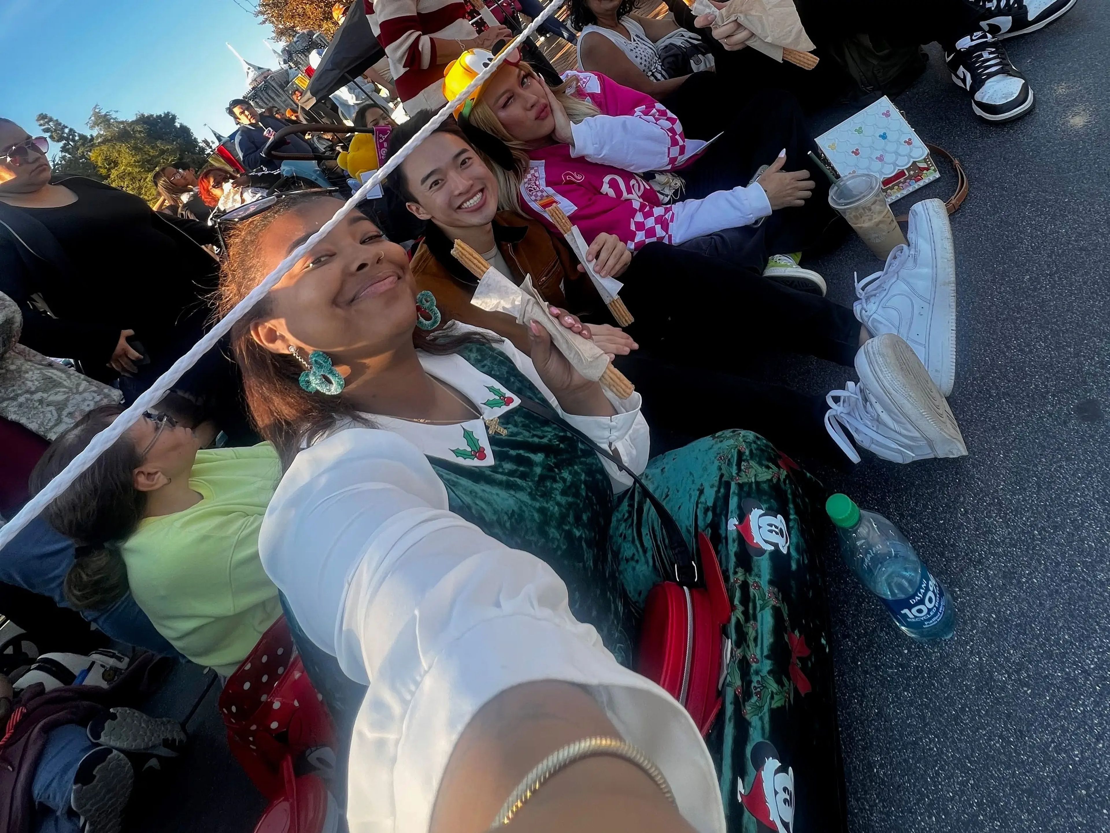 Selfie of the writer wearing a green dress with holly details and two friends, wearing a brown jacket and a pink outfit, sitting on the ground at Disneyland