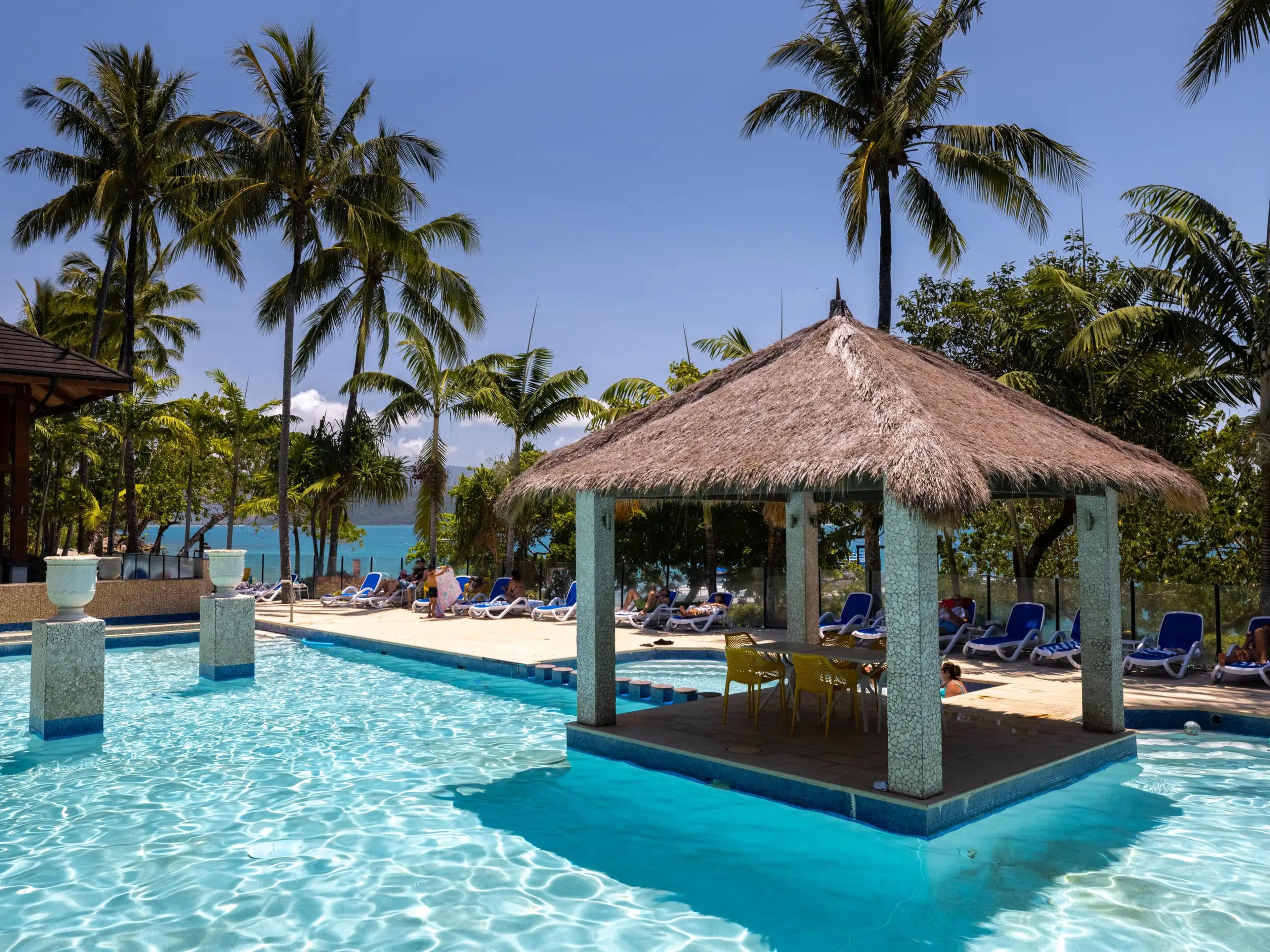 A tropical pool surrounded by palm trees. In the pool is a hut with a platform that houses a table and chairs.