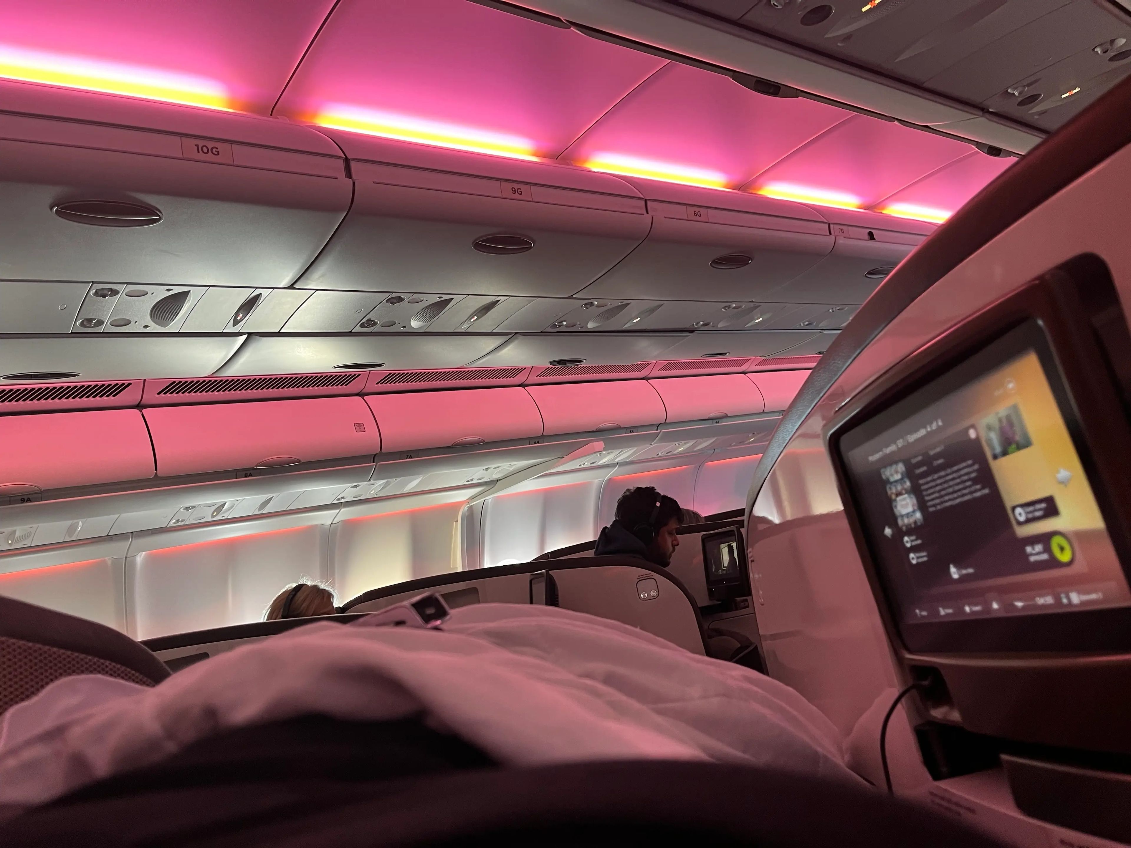 View from the writer laying flat on seat converted into bed on Virgin Atlantic flight. She can see other passengers in their seats and a screen