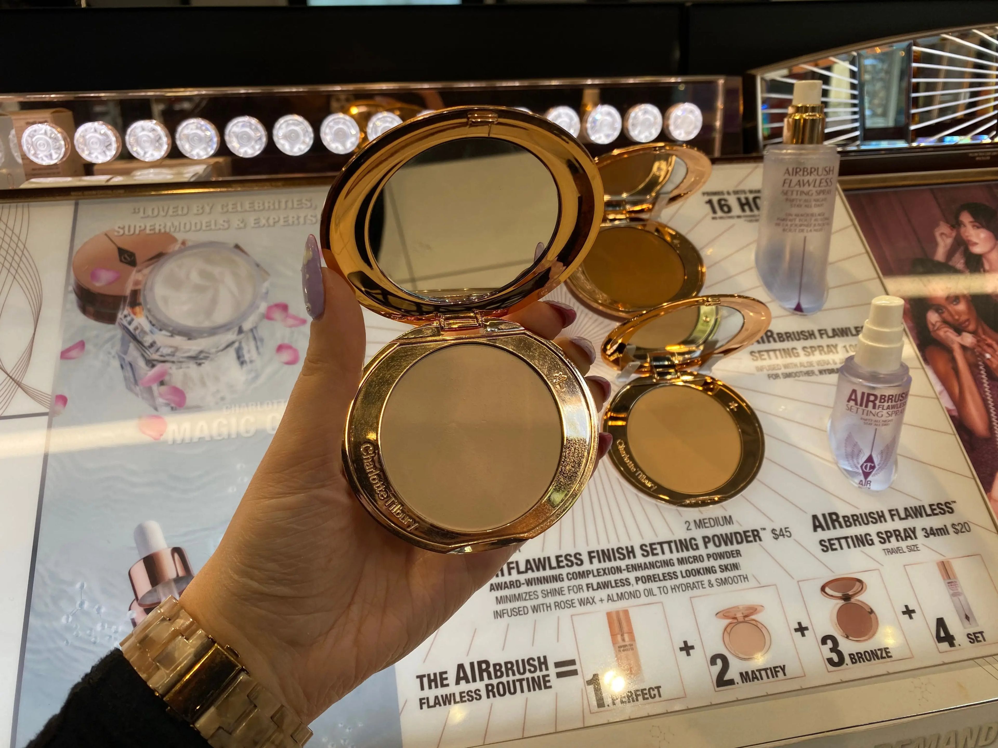 The writer holds a Charlotte Tilbury powder open in front of a display with blue imagery and bright lights in Sephora. The reflection of a white-tile floor can be seen in the mirror of the powder compact.