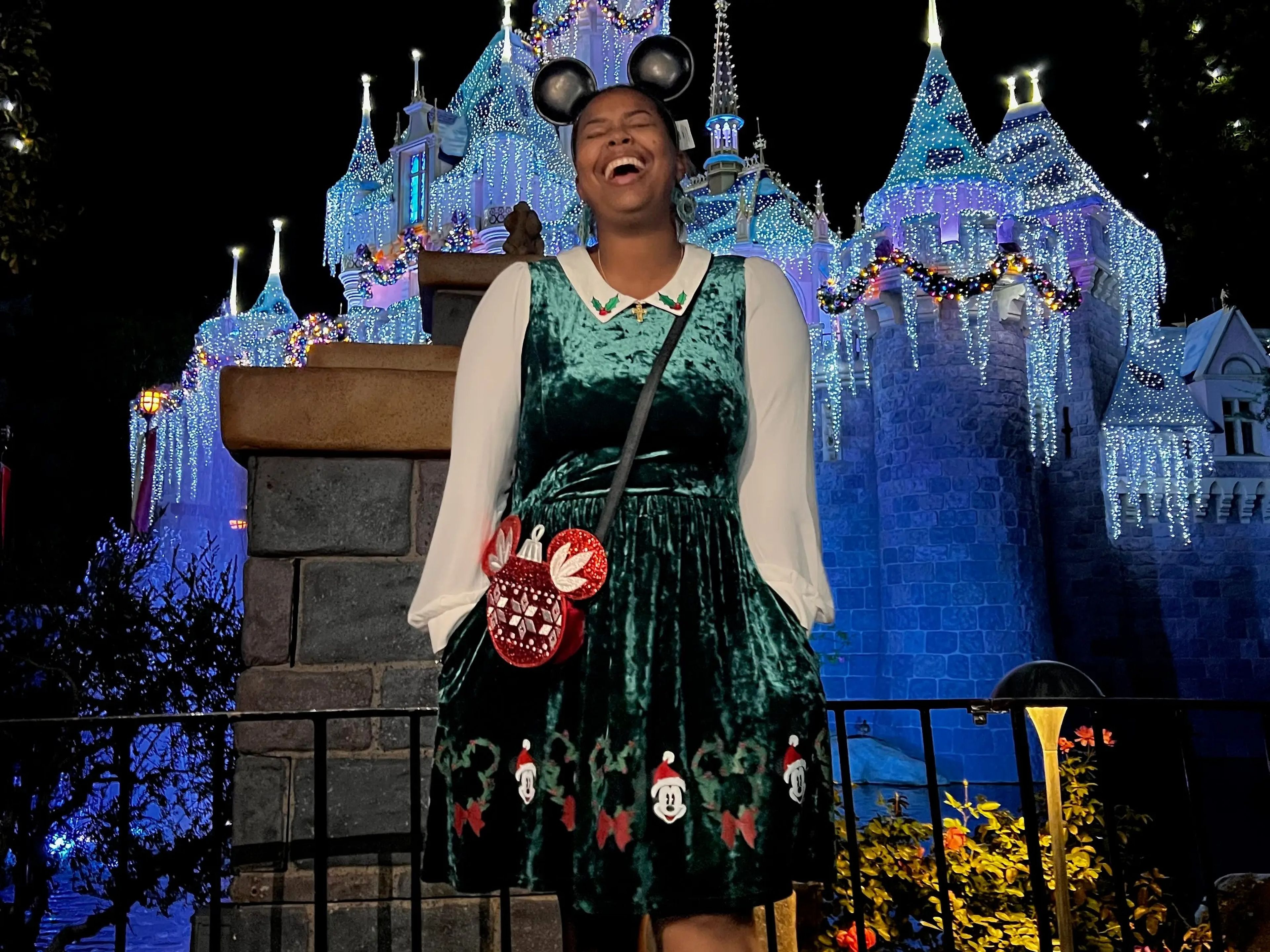 The writer wears a green dress with a Mickey mouse pattern along the hem and mouse ears and stands in front of the castle at Disneyland