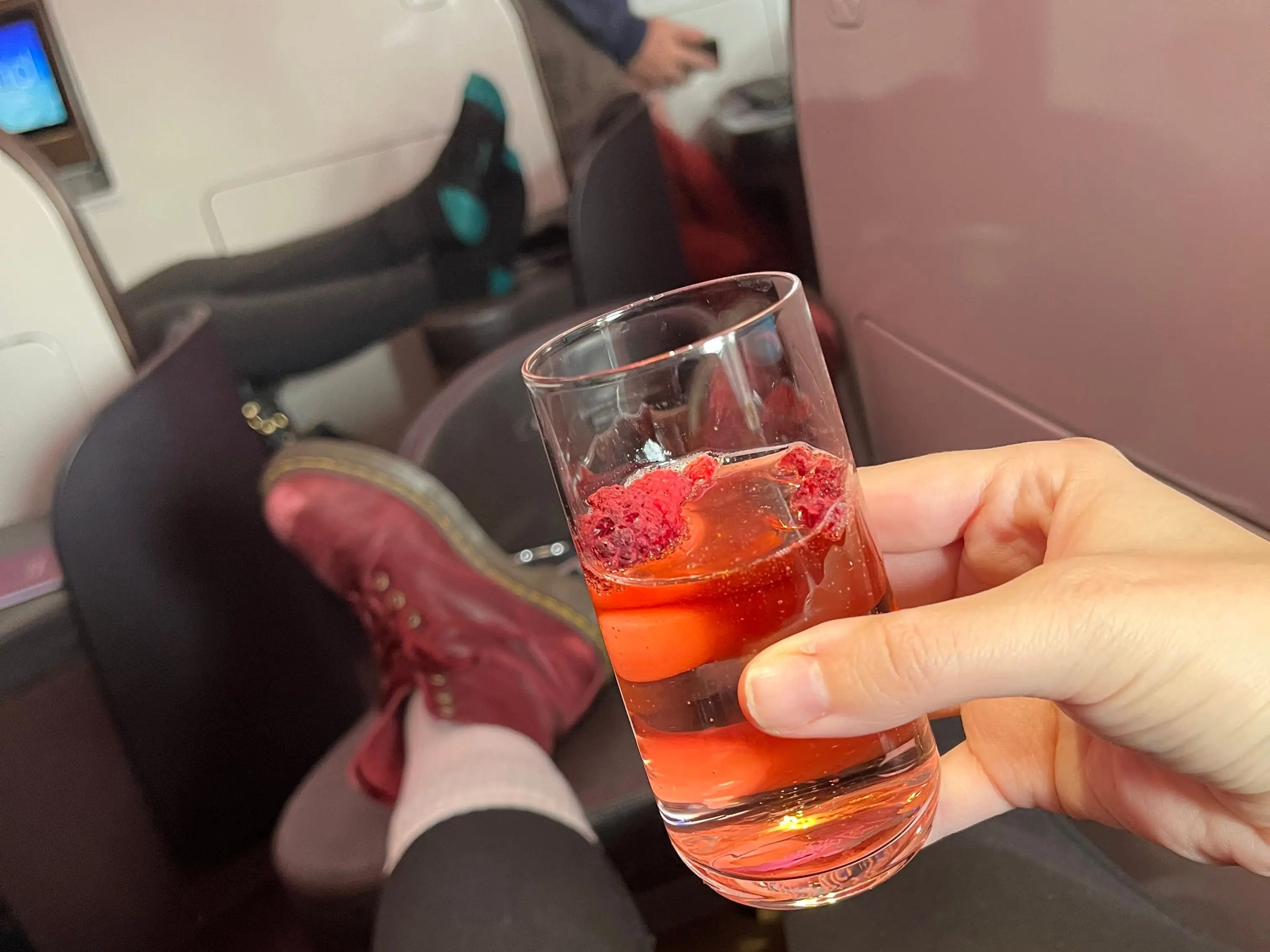 The writer's feet sit on a footrest and point toward other passenger's seats. A hand holds a red-orange cocktail with raspberries floating on top