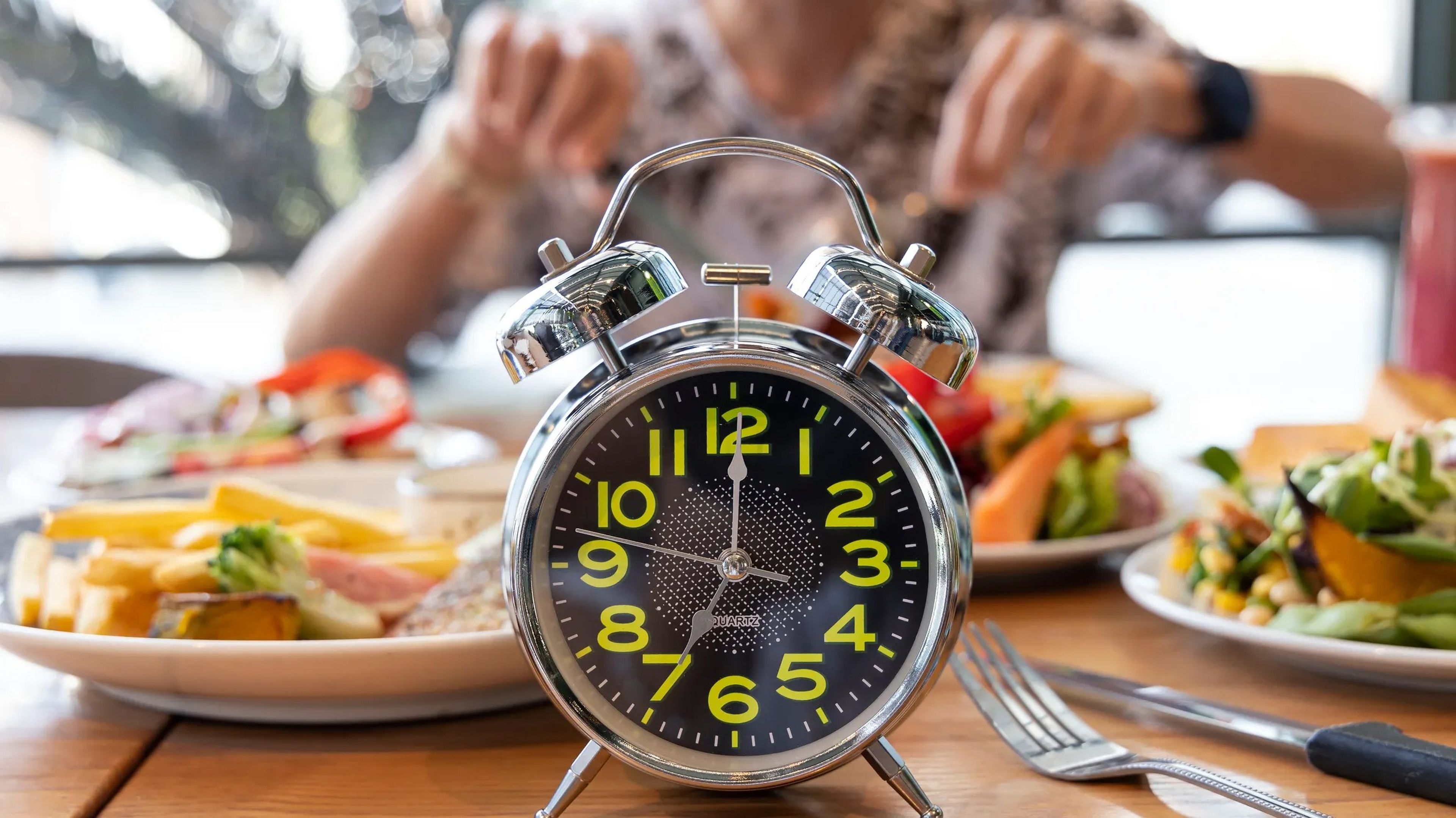 An alarm clock in front of a dinner table full of food with a person in the background preparing to eat on an intermittent fasting schedule.