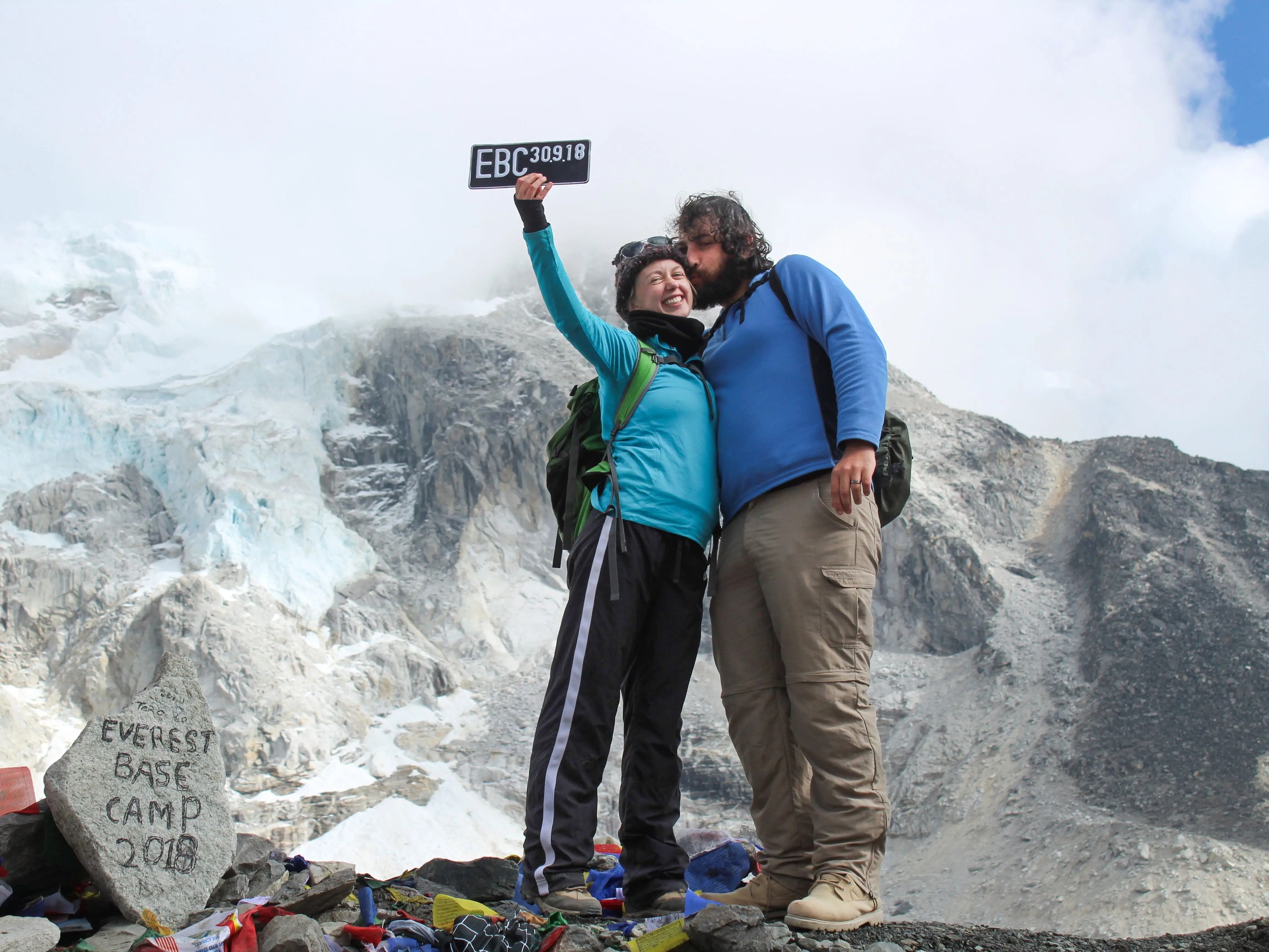 Alex's husband kisses her cheek on the top of a mountain while she holds a sign that reads "EBC 30.9.19." The rock next to them has an inscription that reads "Everest Base Camp 2018."