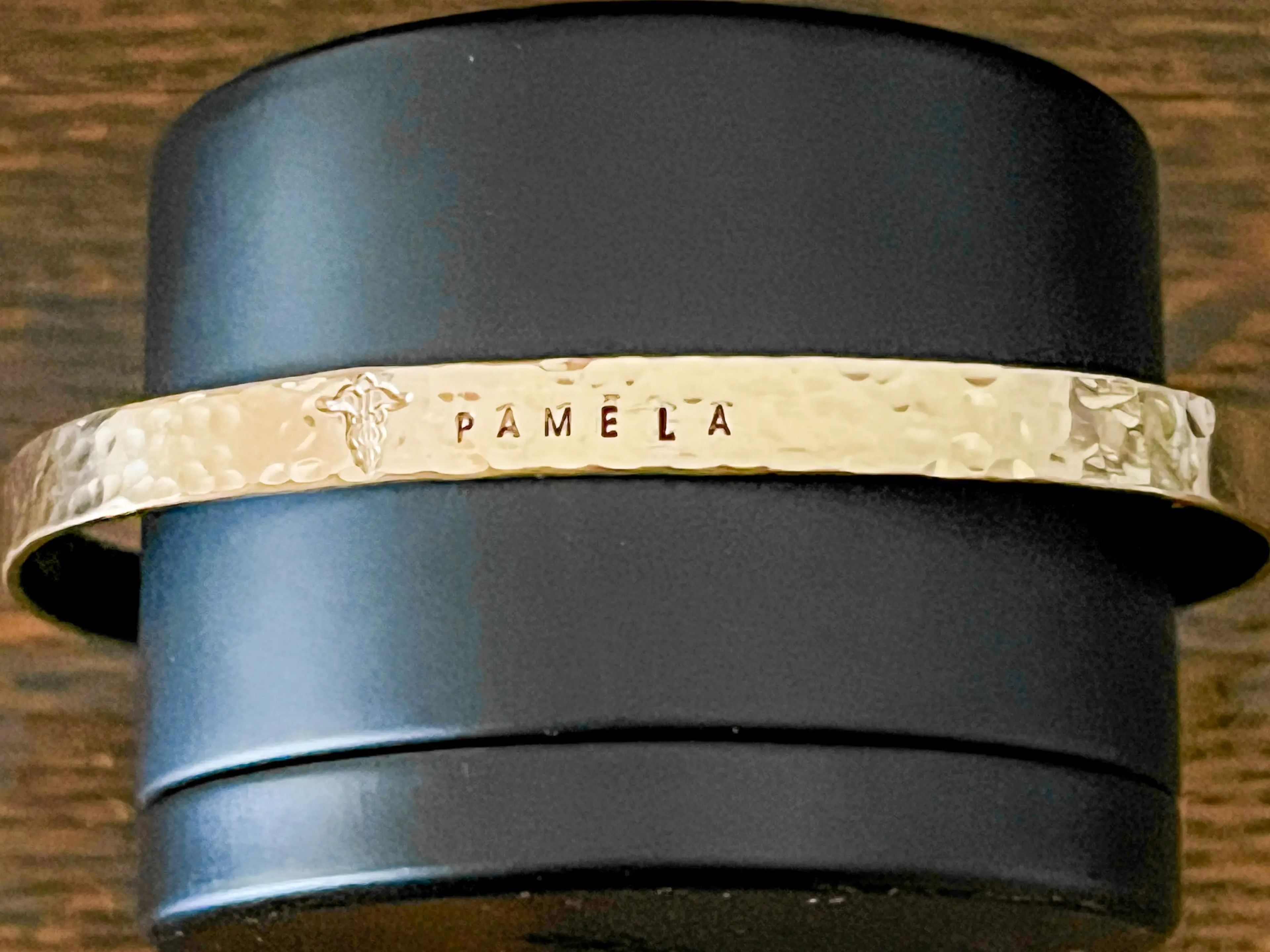 A black bracelet holder on a wood floor showcases a gold bracelet with the name "Pamela" engraved on it