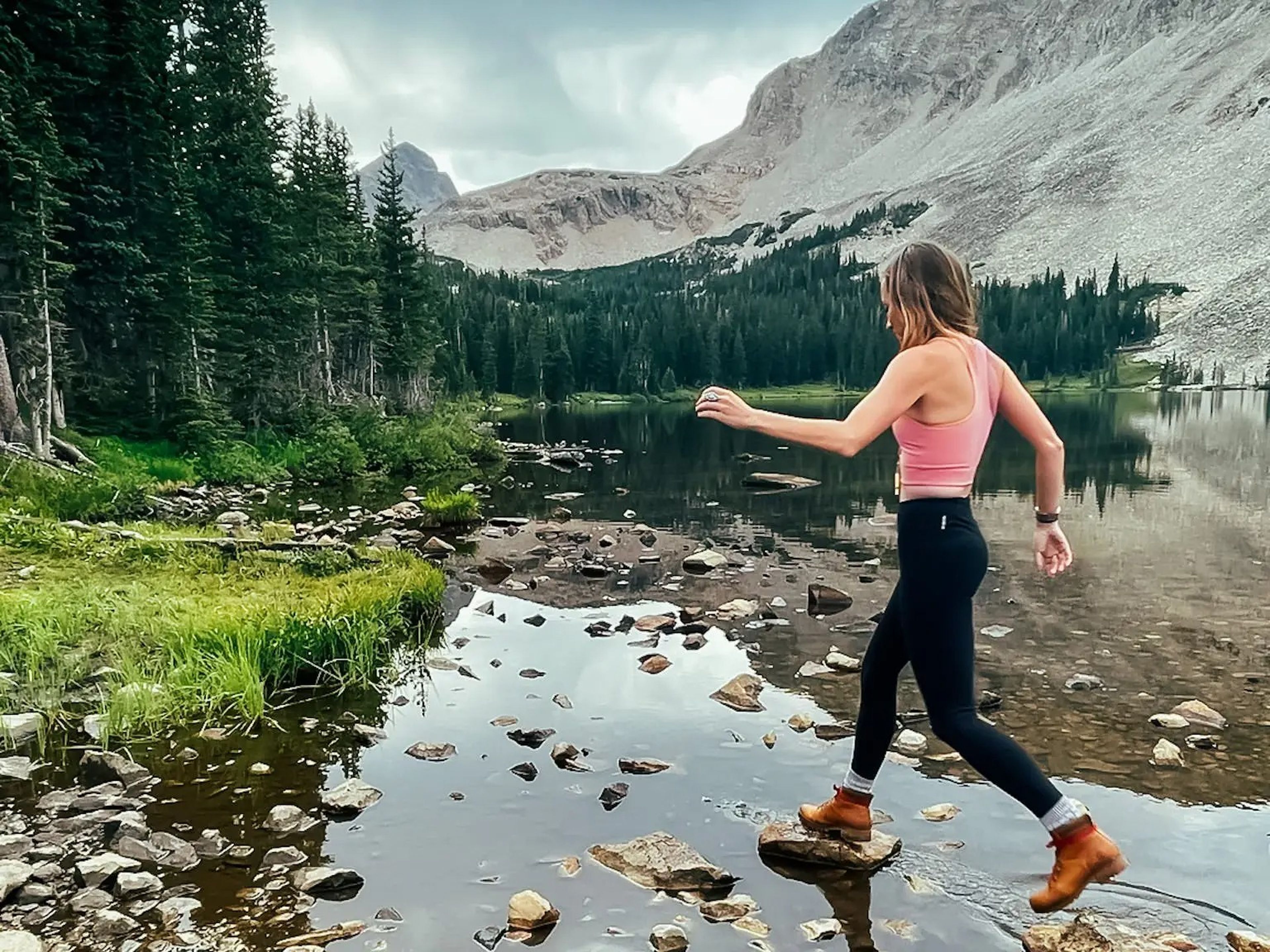 Emily, wearing a pink tank top, black leggings, and boots, walks across the rocks in a lake. Behind her are tall trees and mountains.