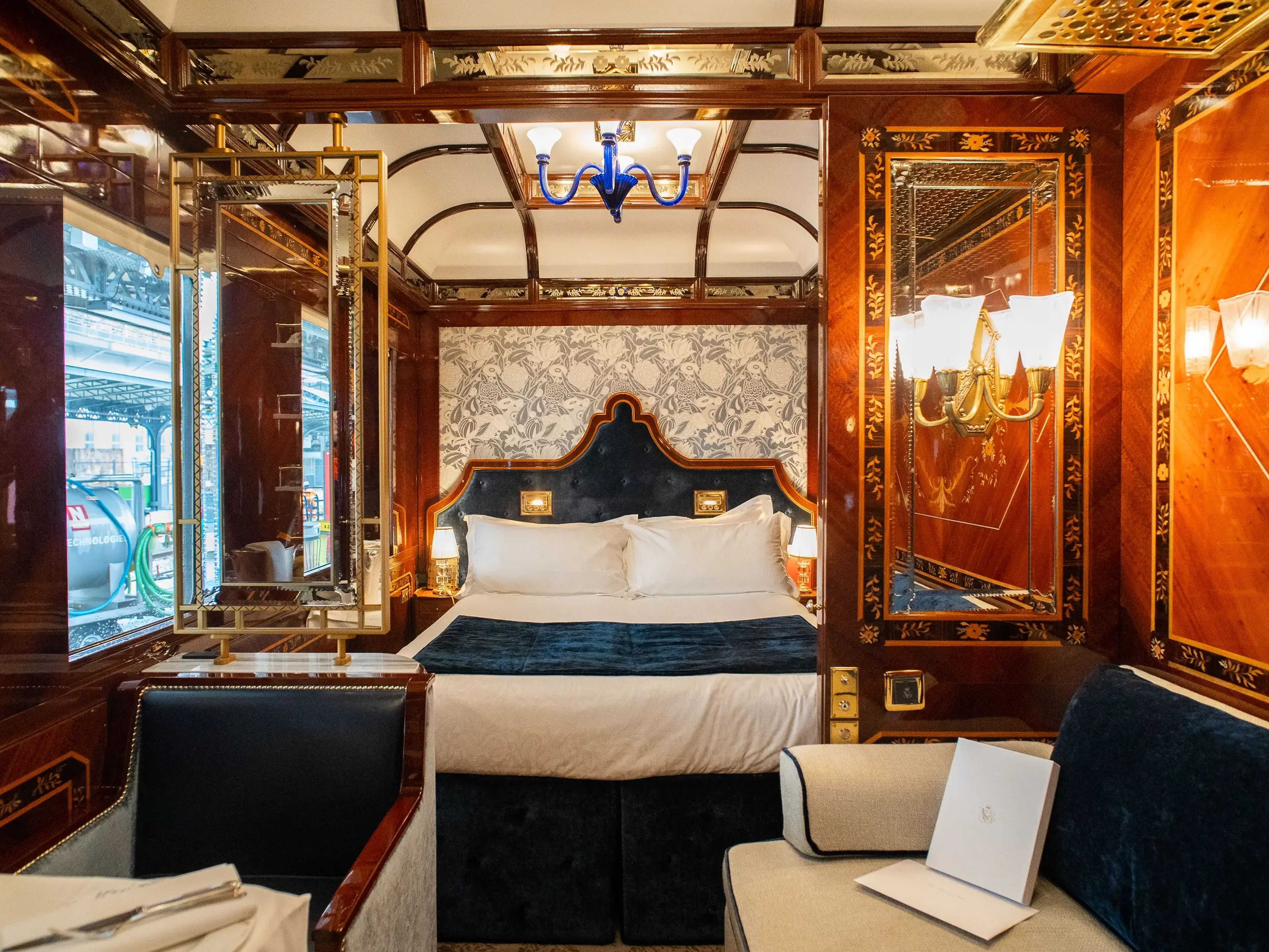 Inside a wood-walled train suite with white and navy blue furnishings, including a seat on the left, a couch on the right, and a bed in the back center.