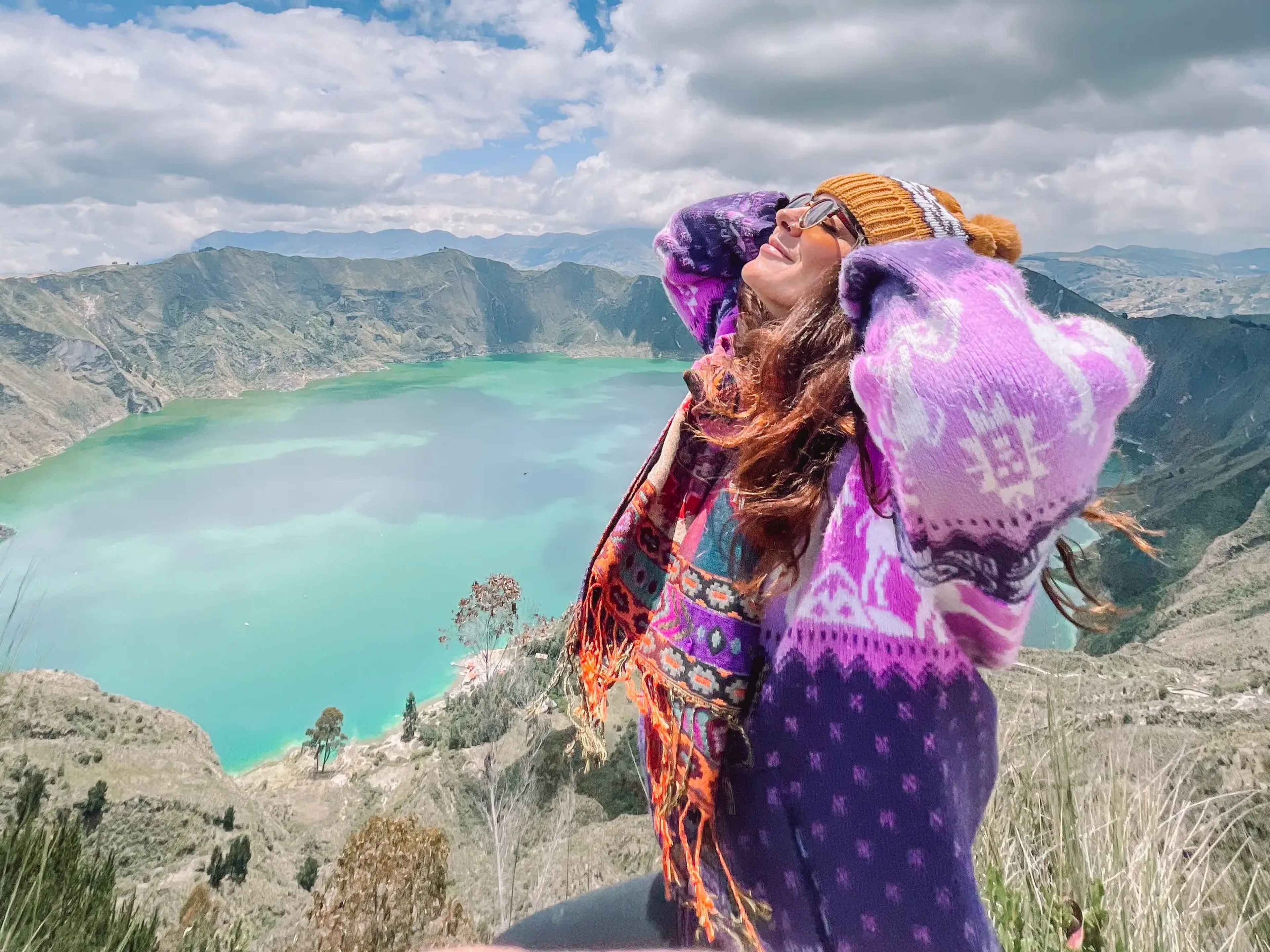 Kate Boardman wears a pink and purple sweater, sunglasses, and a colorful scarf, while standing at the rim of a cliff looking down to a body of water.
