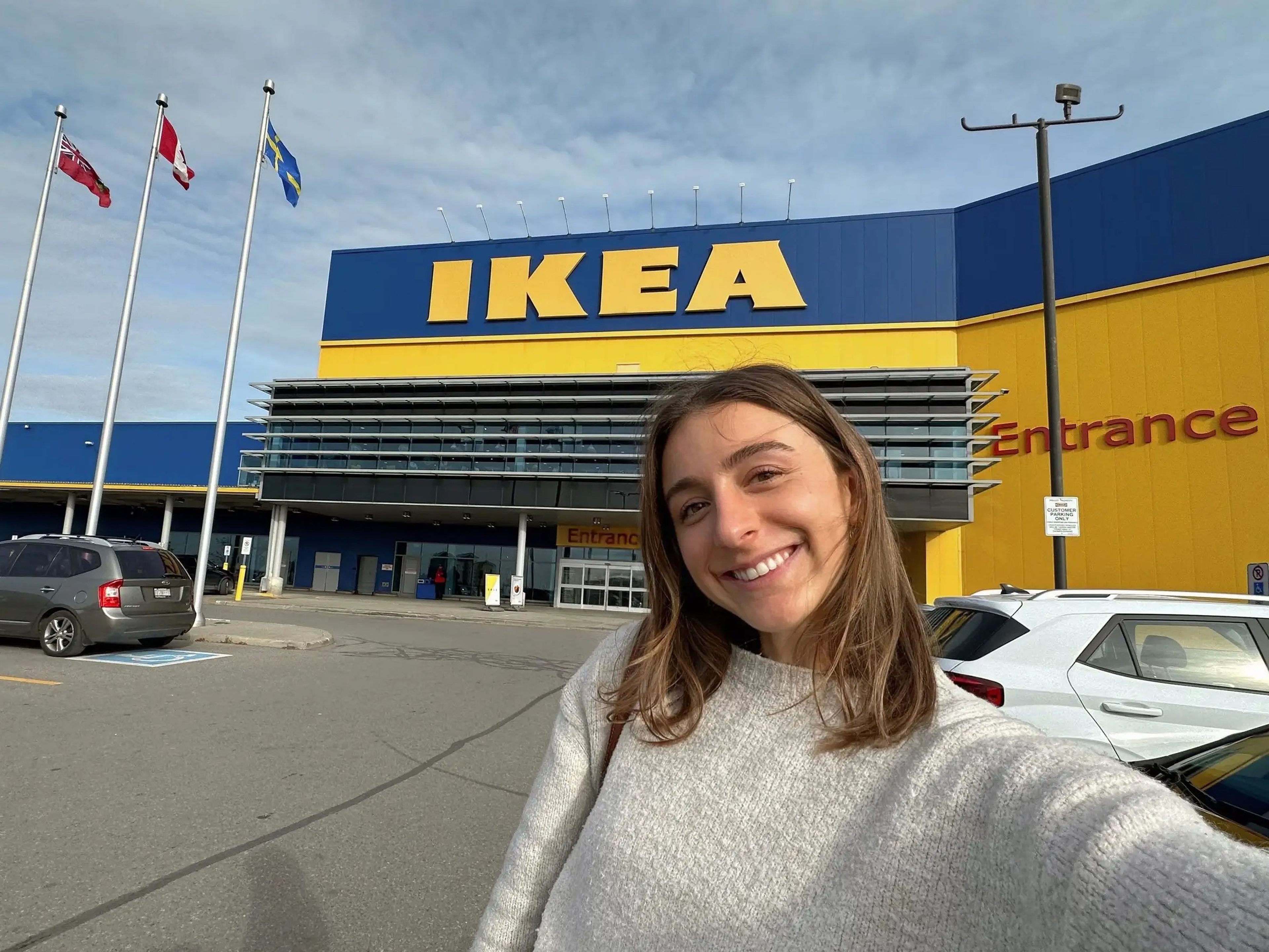 Selfie of the writer smiling and wearing a white sweater in Ikea parking lot in front of the store