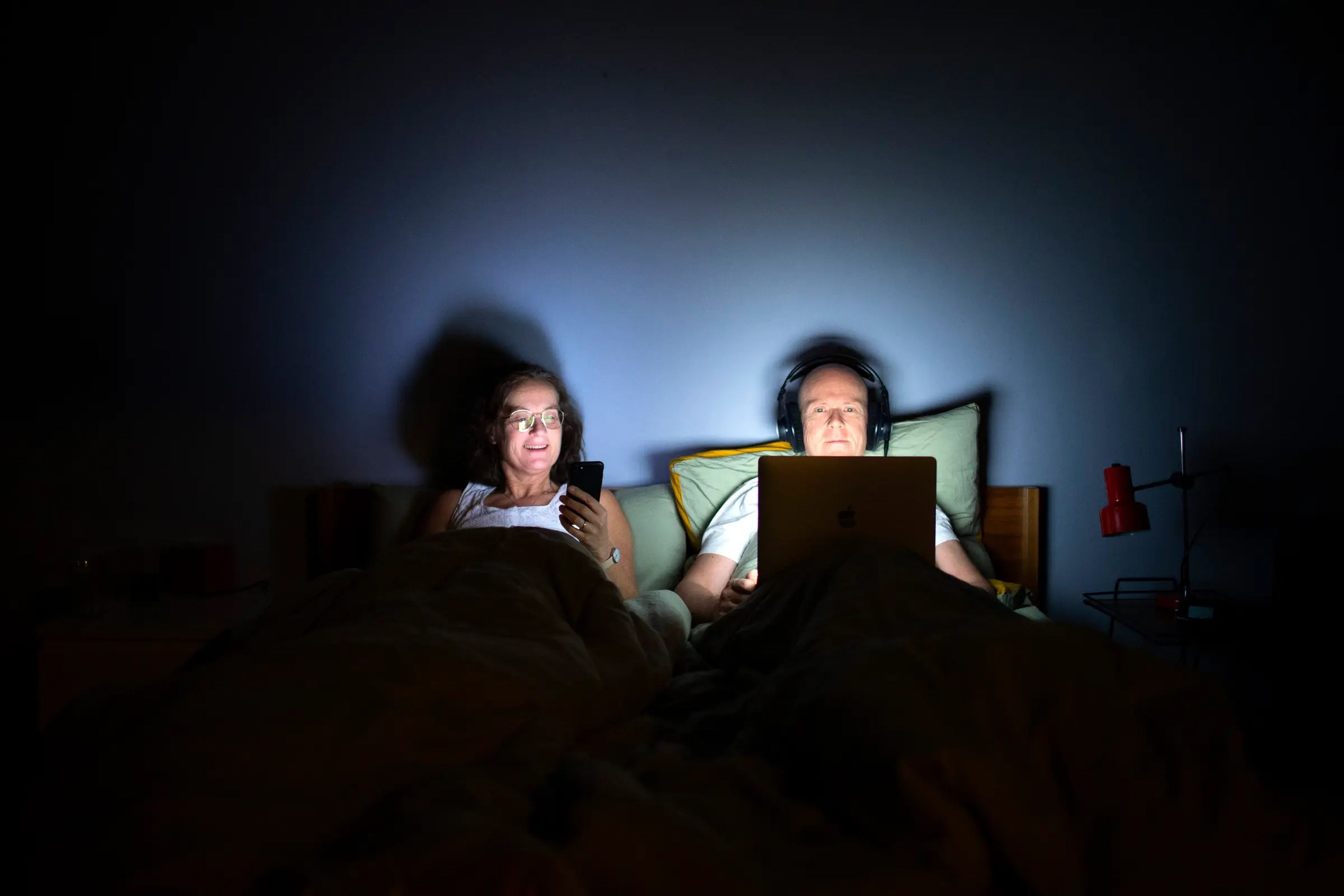 A woman looks at her phone, and a man watches his laptop in bed at night.