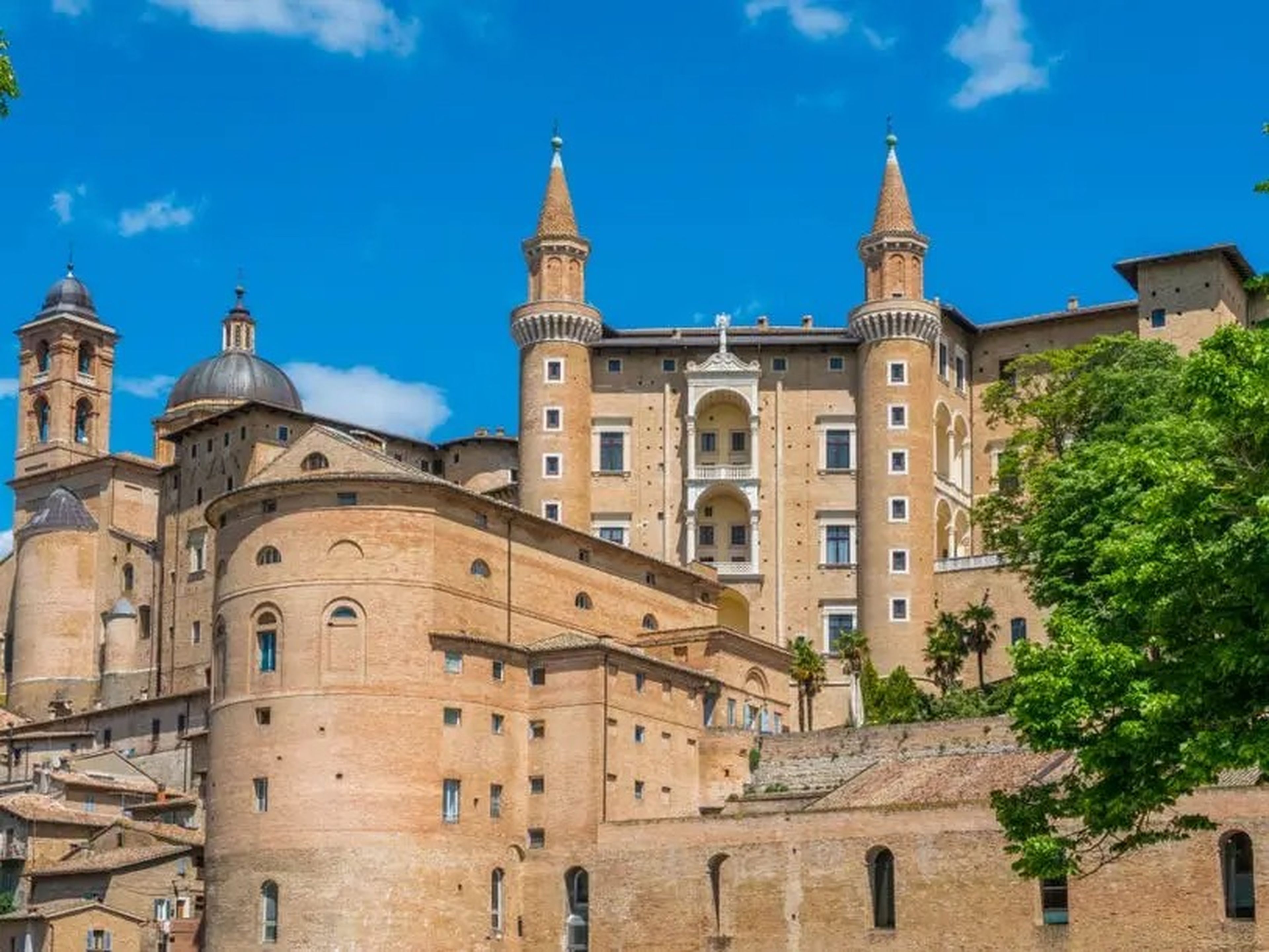 Buildings that look like castles in Urbino, Italy. The sky is blue, and there are trees on each side of the building.