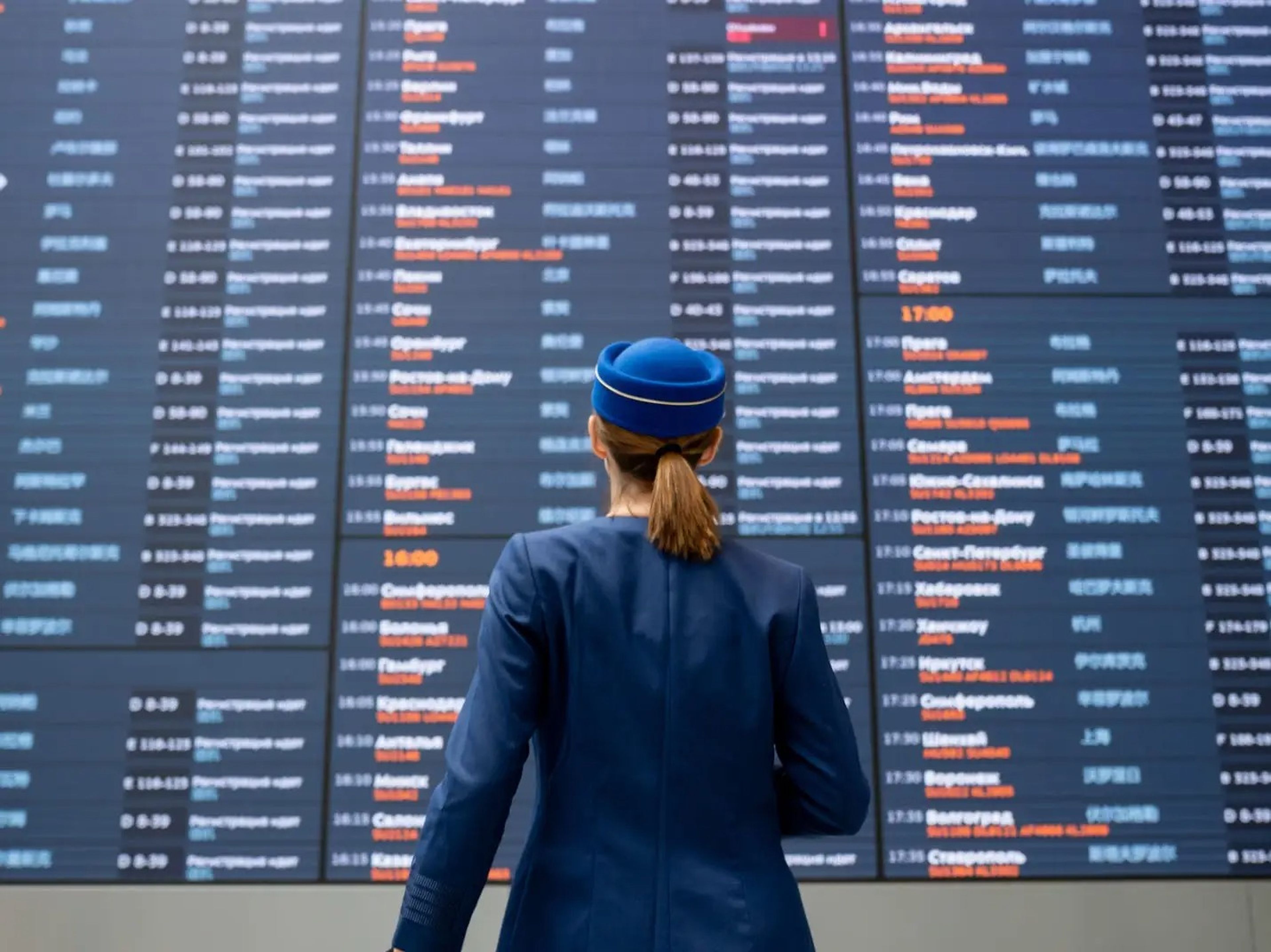 A flight attendant with a blue hat and blazer looking at a wall of flight schedules at an airport