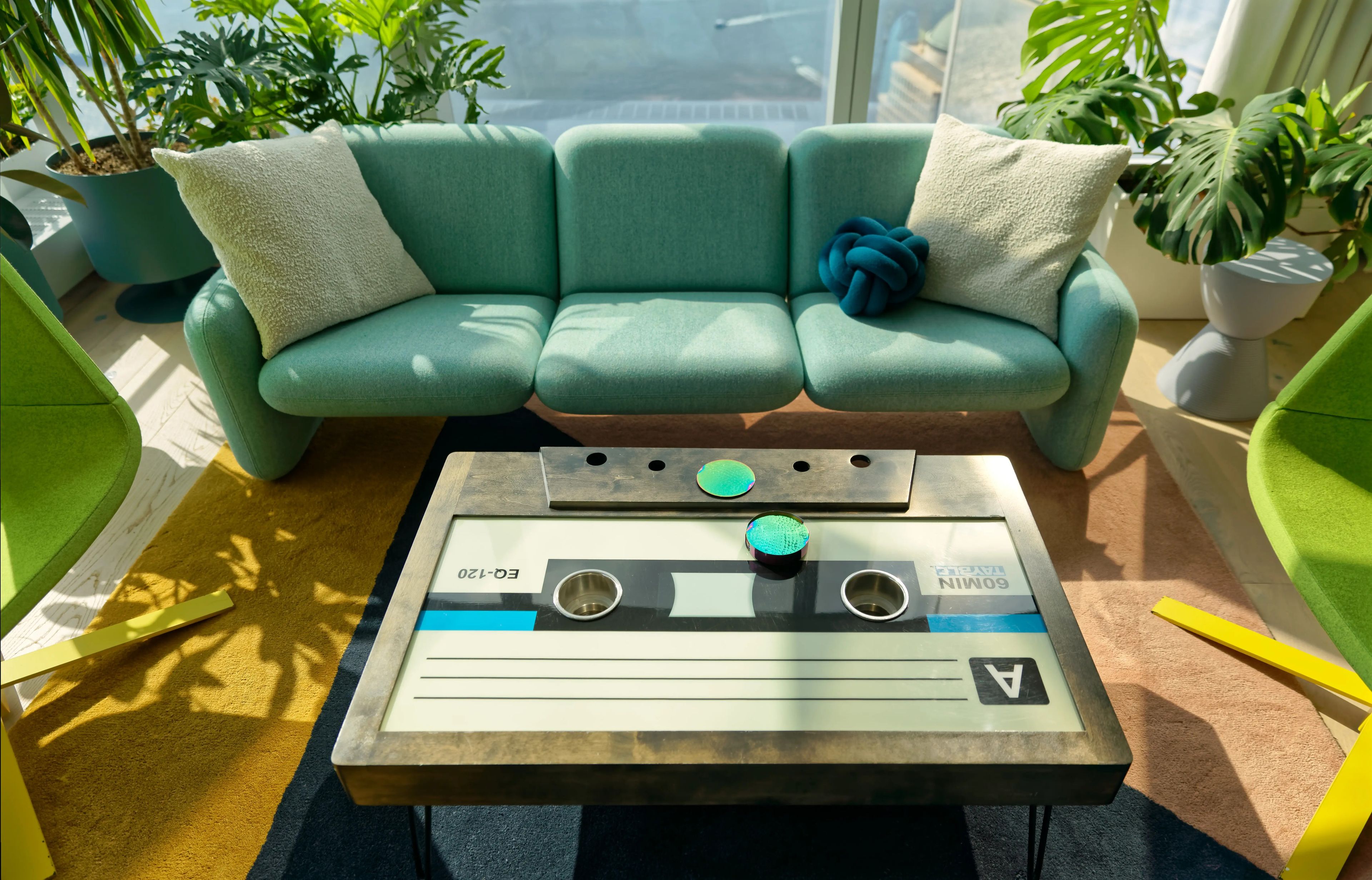A green couch with a coffee table that is shaped and designed to look like a cassette tape. The room has a lot of natural light and a few plants in it.